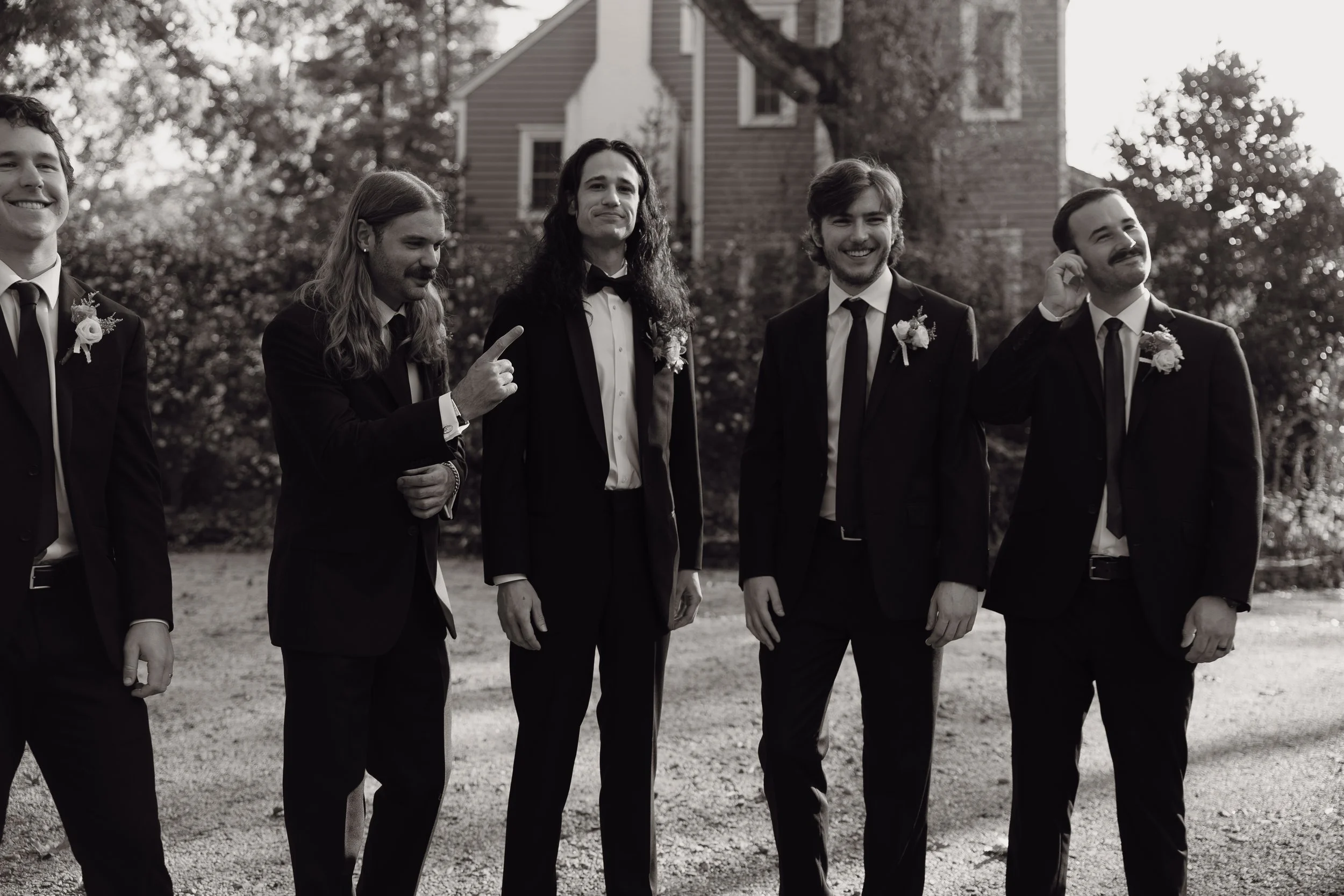 Group of five men in suits standing outdoors, some with boutonnières, smiling and posing in front of a house and trees.