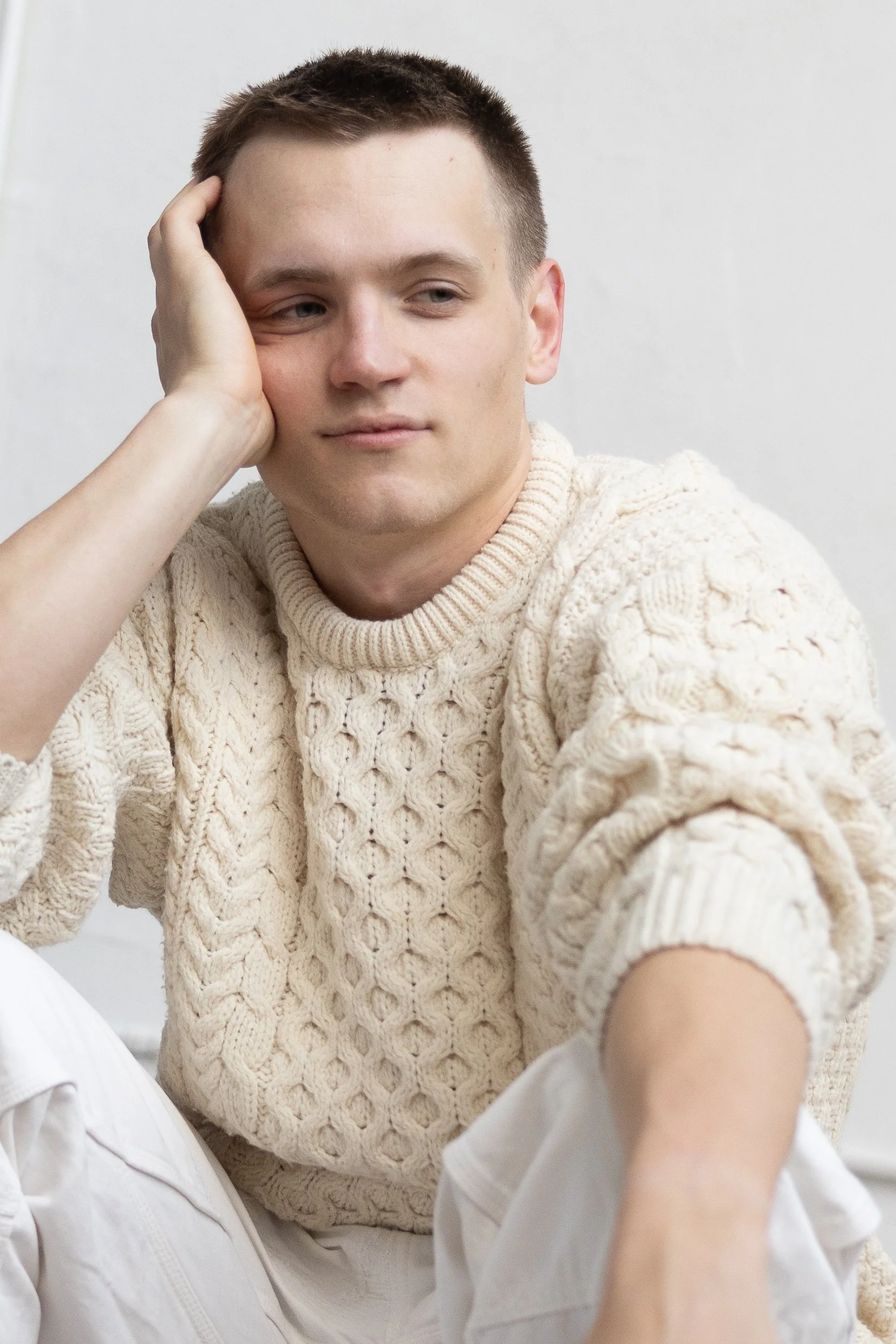 Young man sitting with his hand resting on his head, wearing a cream-colored cable knit sweater, with a thoughtful expression.