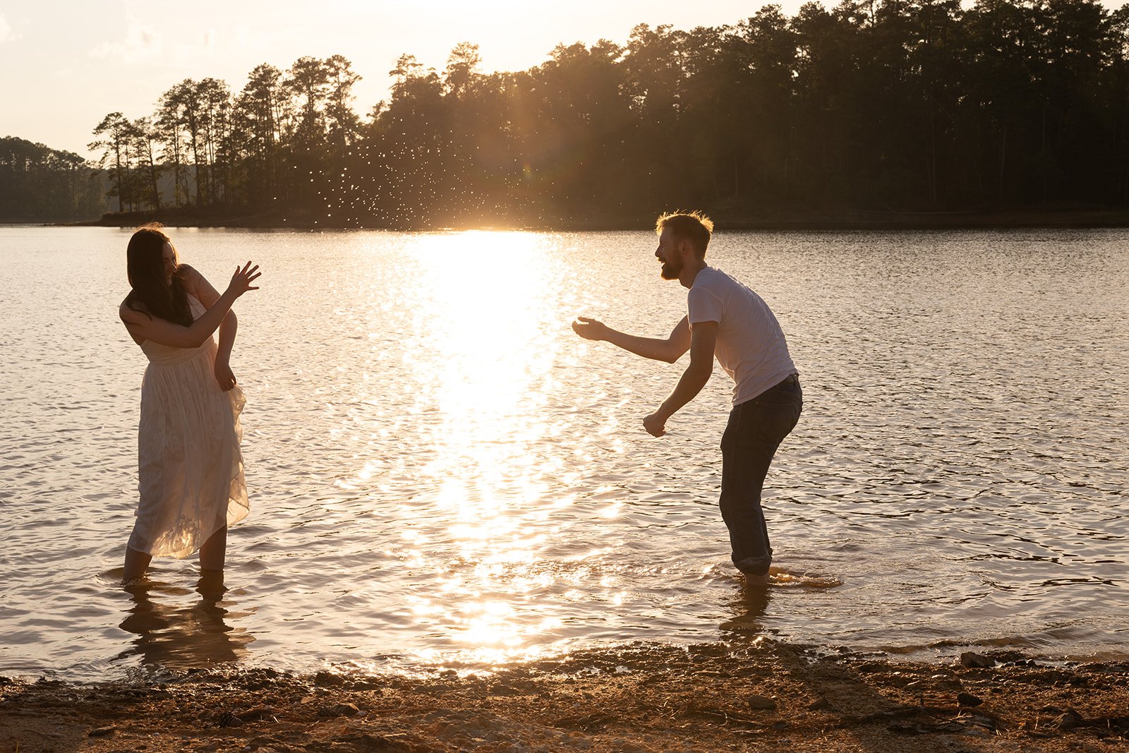 A man and woman playing and splashing in the water at sunset, near a lakeshore with trees in the background.