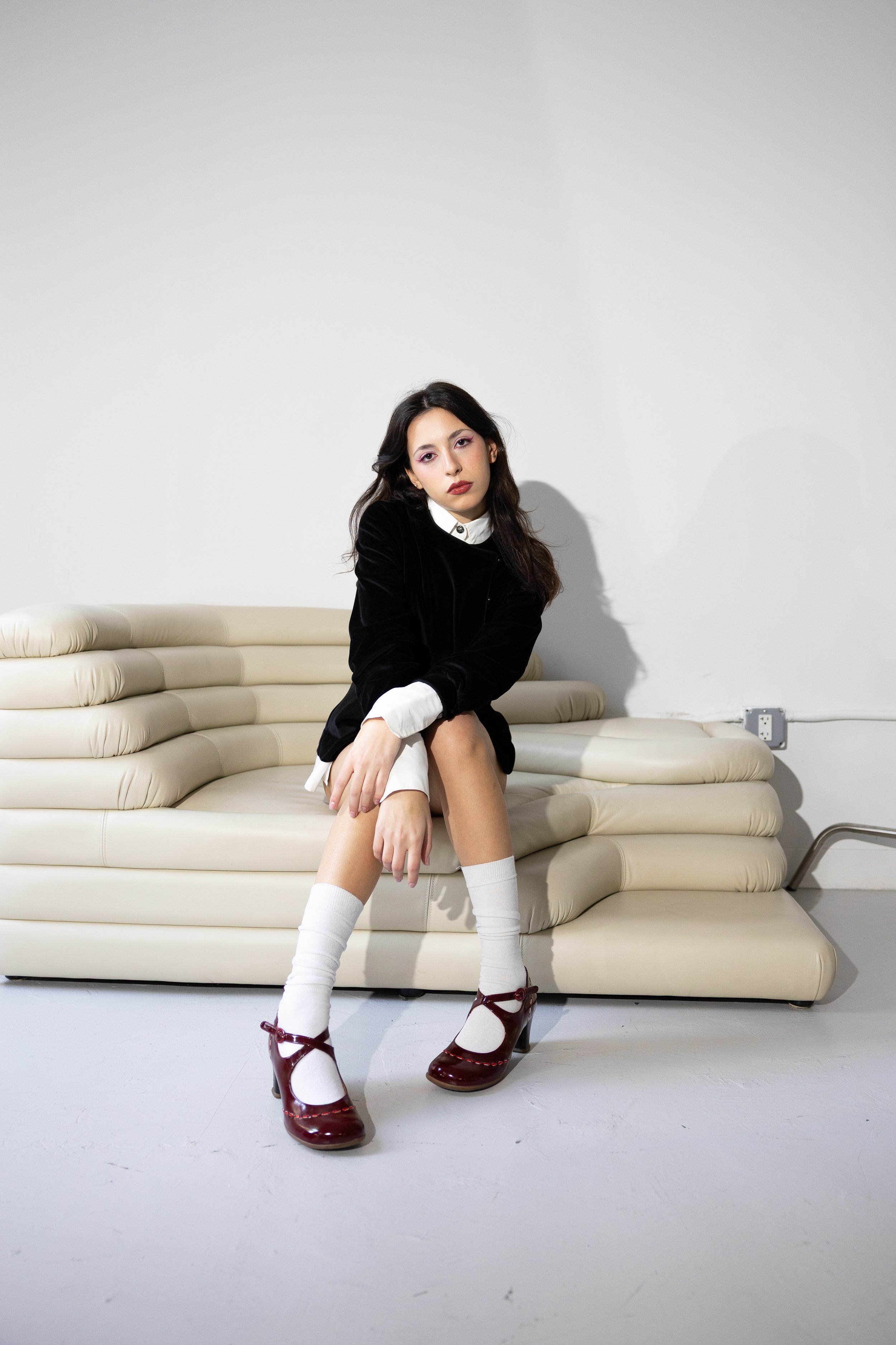 A young woman with long dark hair, wearing a black velvet blazer, white shirt, white knee-high socks, and burgundy patent shoes, sitting on a cream-colored vintage sofa in a room with white walls and visible electrical outlets.