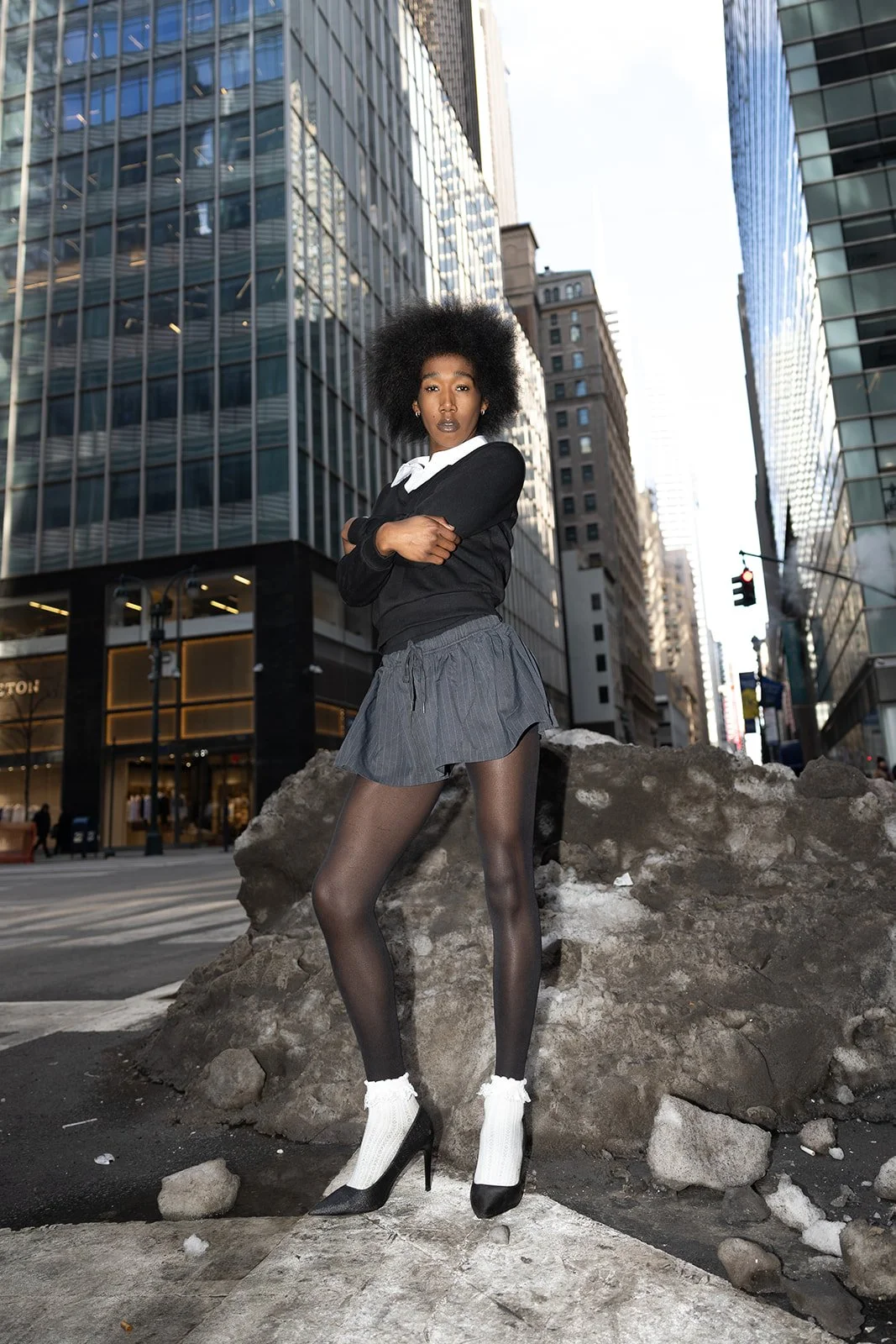 A woman with afro hair stands on a city street in front of large rocks, with tall glass buildings in the background. She is wearing a black sweater, a white collared shirt, a short gray skirt, sheer black tights, and high heels with white socks.