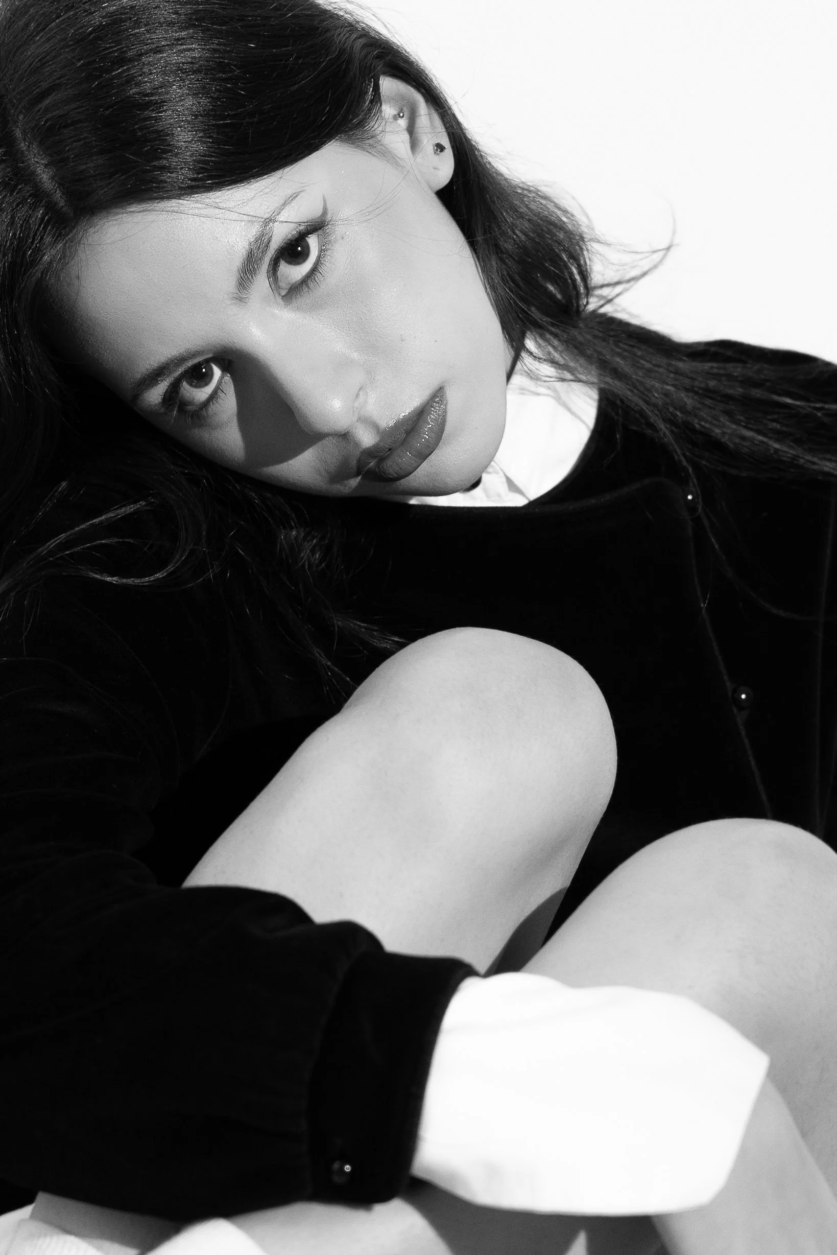 Black-and-white portrait of a young woman with dark hair, piercing eyes, and glossy lips, wearing a dark top and white shirt, sitting against a plain background.