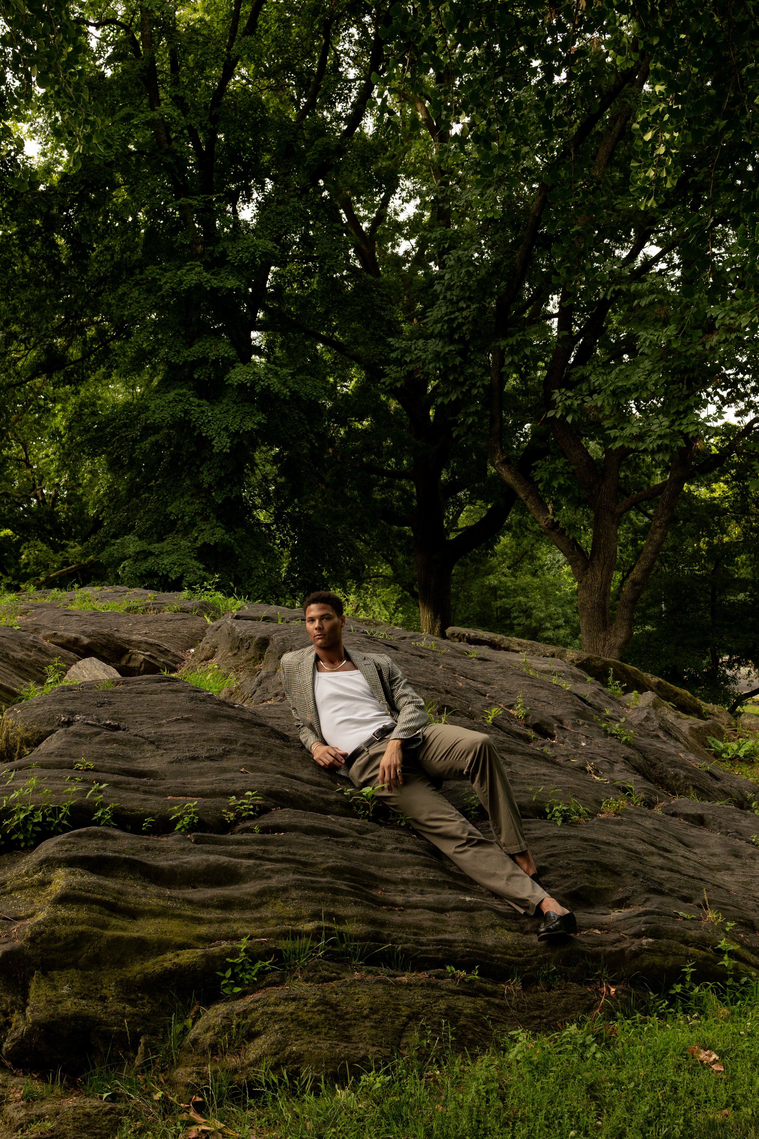 A man in a patterned blazer and khaki pants reclining on large rocks outdoors with green trees in the background.