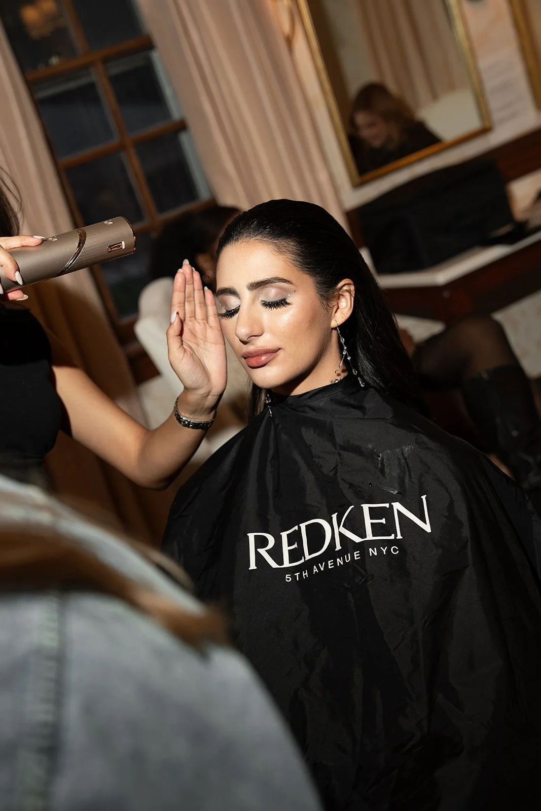 A woman with dark hair and makeup getting her hair styled at a salon, wearing a black cape with the Redken logo in white, inside a room with a window and mirror.
