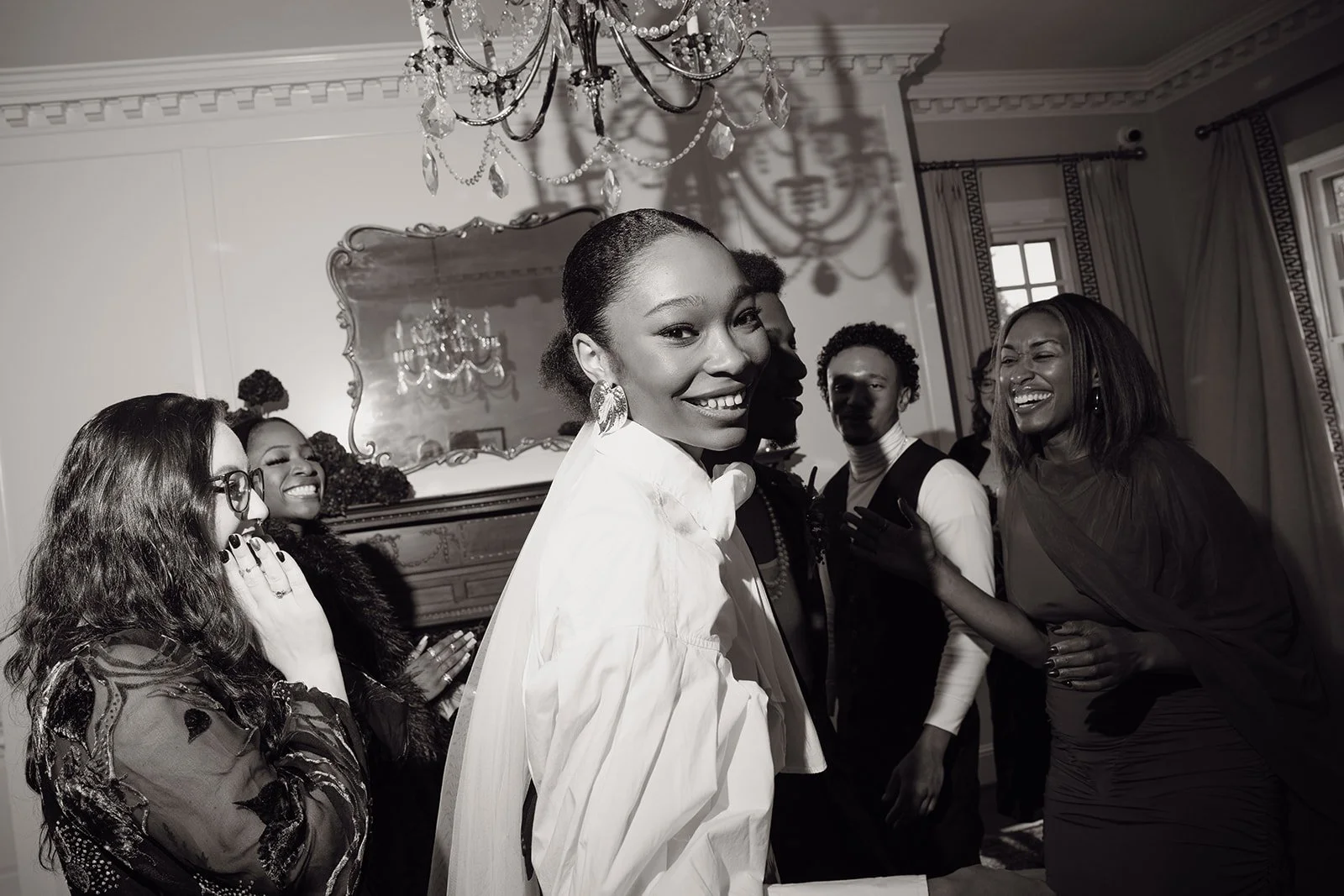 A group of women smiling and enjoying each other's company in an elegant room with a chandelier and a mirror.