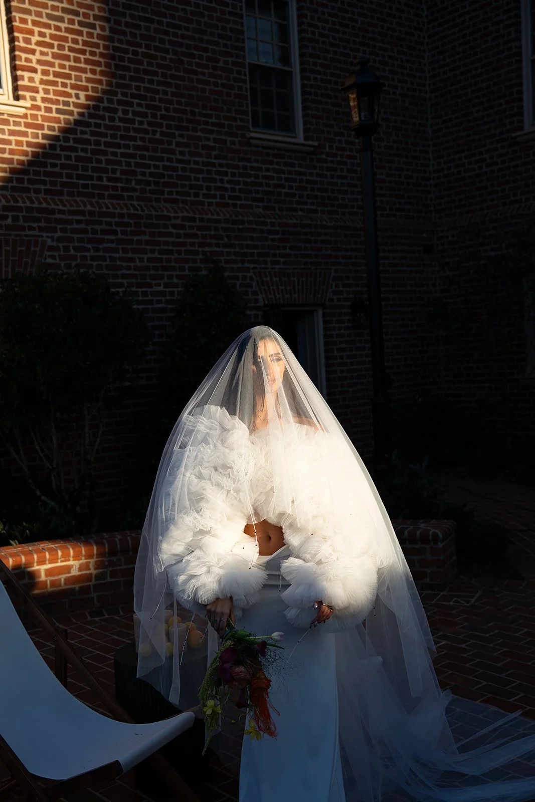 A woman dressed in a white, fluffy garment with a long veil sitting outdoors at sunset, holding a bouquet of flowers, with a brick building, a lamp post, and a chair nearby.