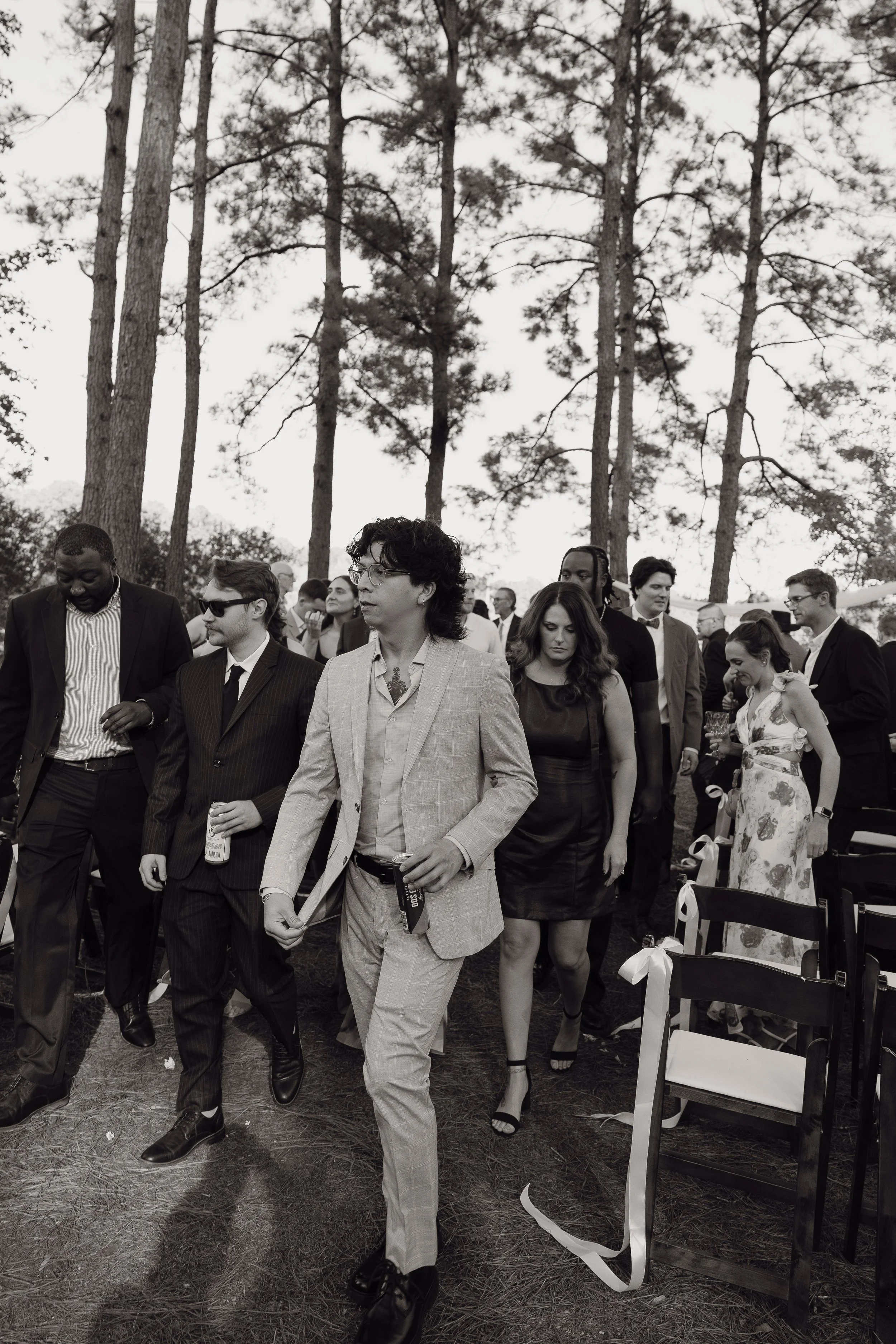 Group of people walking outdoors among trees, dressed in formal and semi-formal attire, some holding drinks.