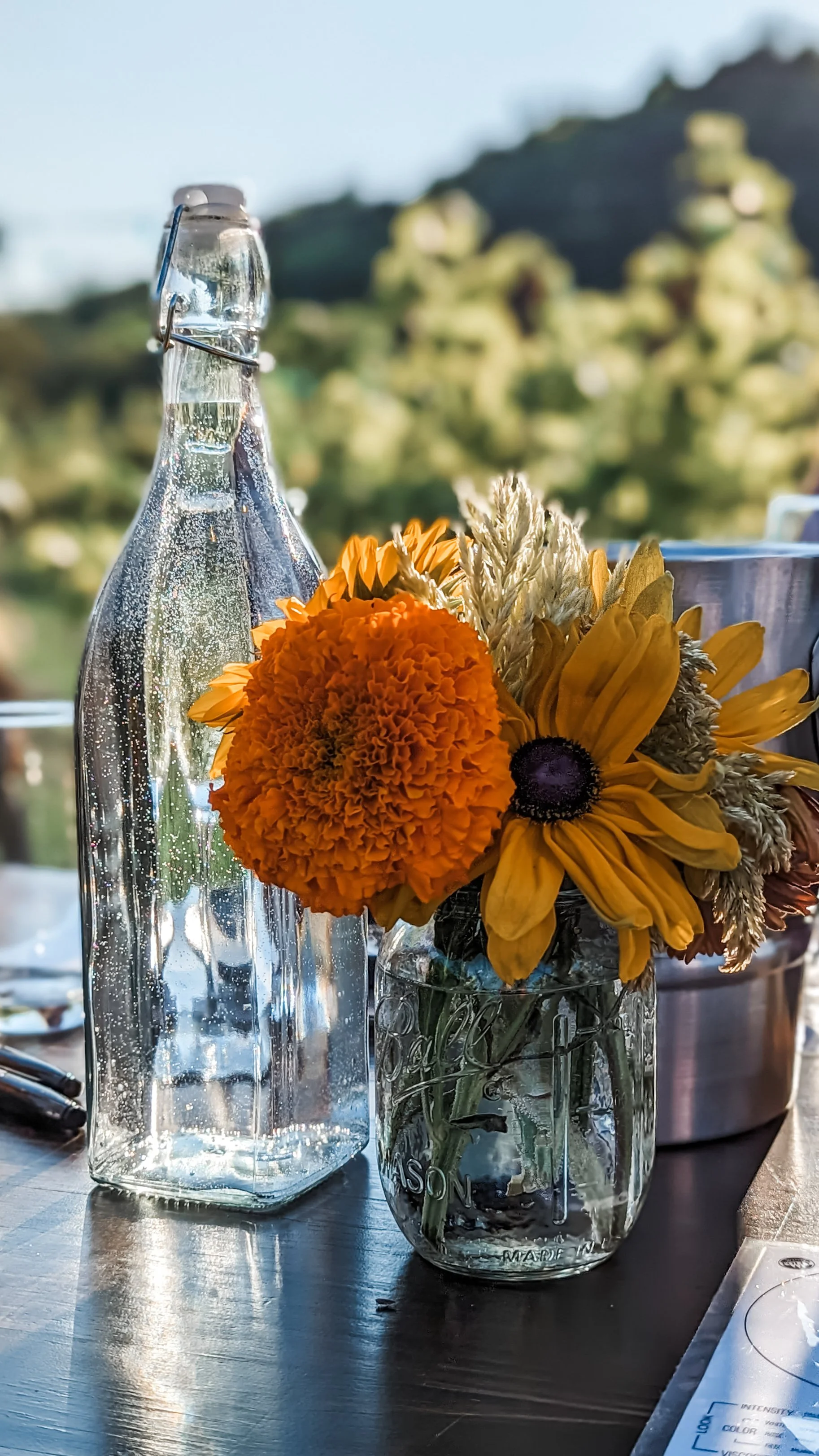 A glass jug with a floral arrangement, featuring orange marigolds and yellow daisies, placed next to a tall empty glass bottle on a wooden table with a scenic hillside background.