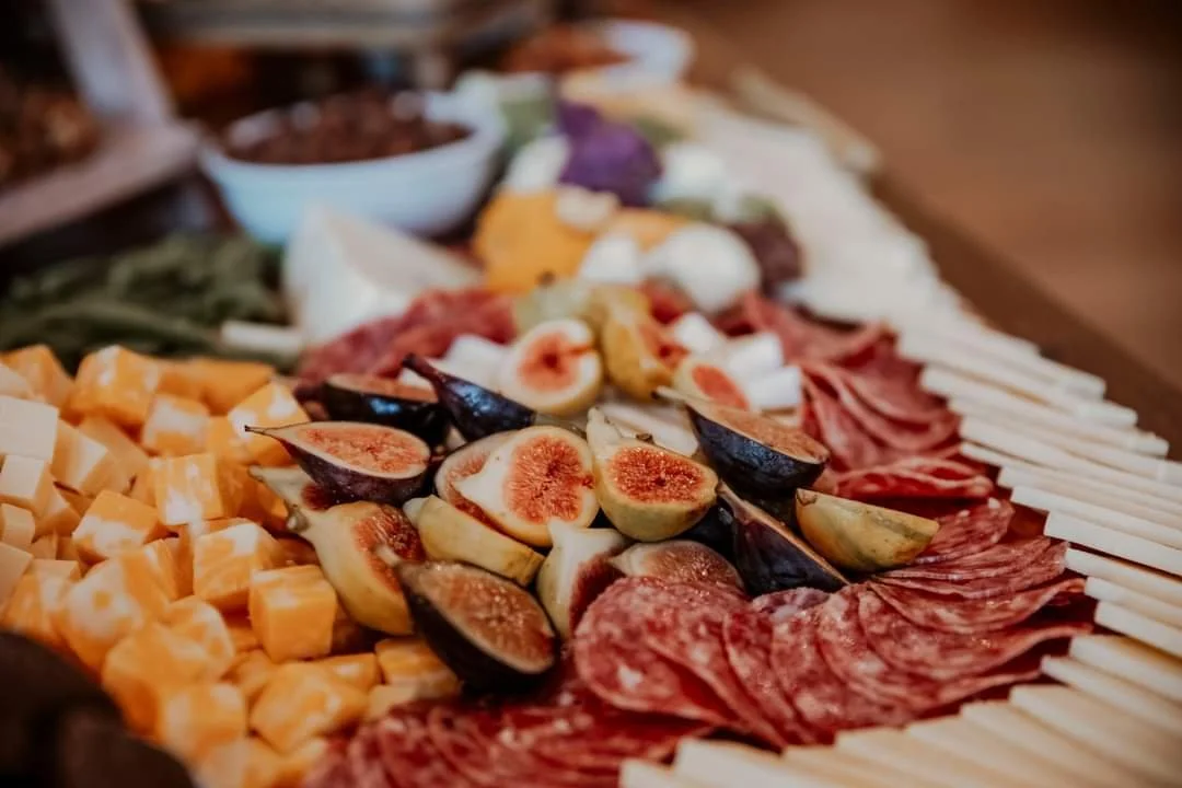 A sliced fig, cheese cubes, cured meats, and cheese sticks on a platter, with bowls of other food in the background.