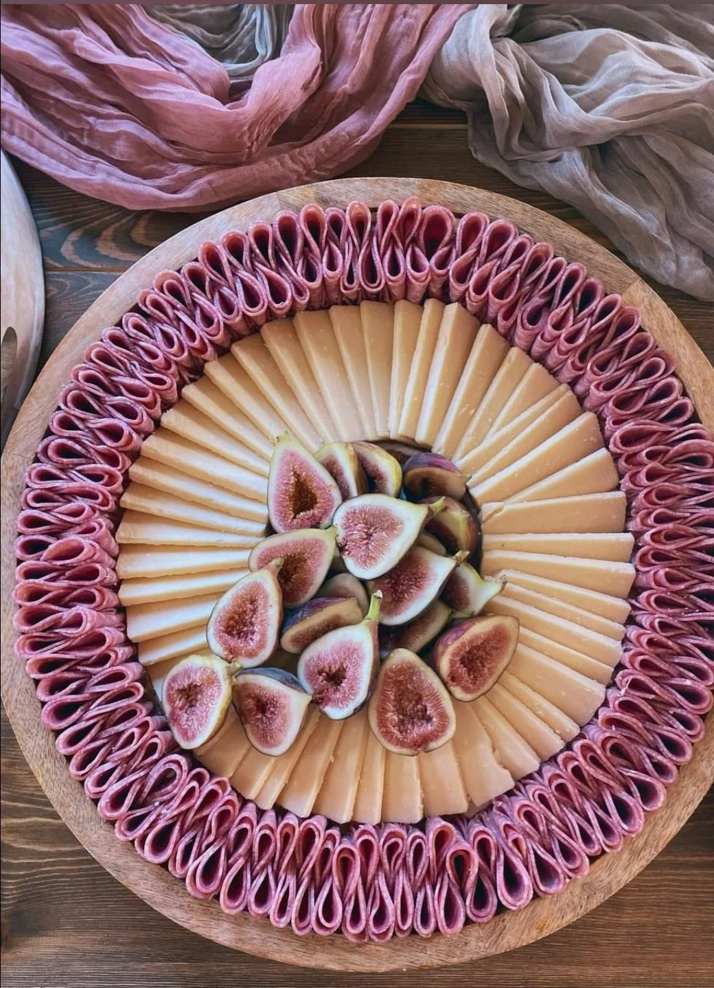 A round wooden platter with sliced cheese arranged in a circle, overlapping slightly. Inside the cheese circle are sliced figs with pink flesh and greenish-purple skin, arranged in a small pile. Pink and beige gauzy fabric is draped at the background.