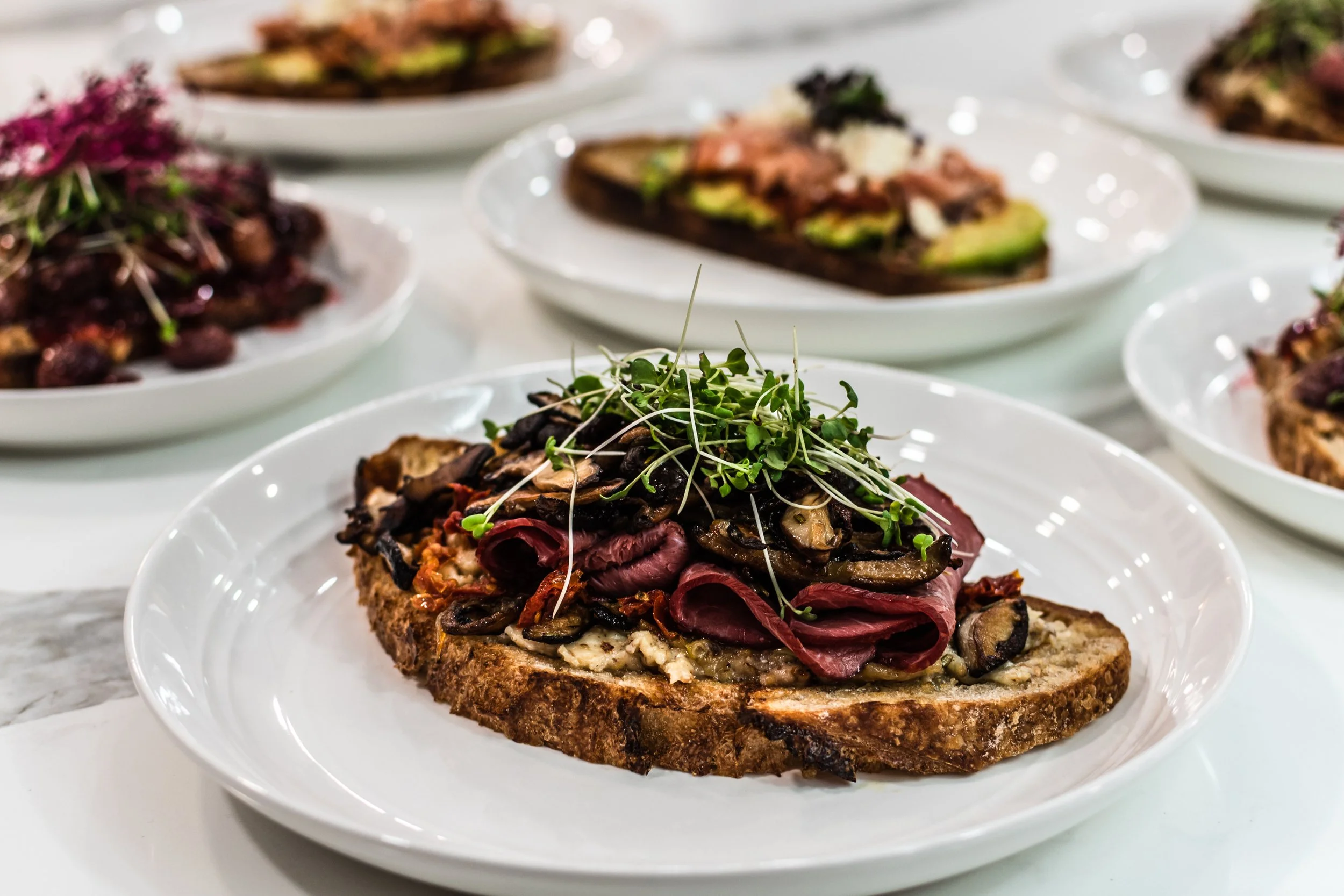 A plate with toast topped with roasted mushrooms, sliced meat, microgreens, and cheese.
