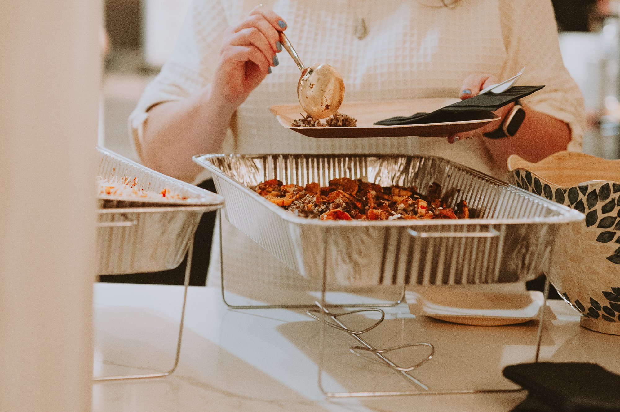 Person serving food from a tray at a buffet table, with food on a plate in hand.