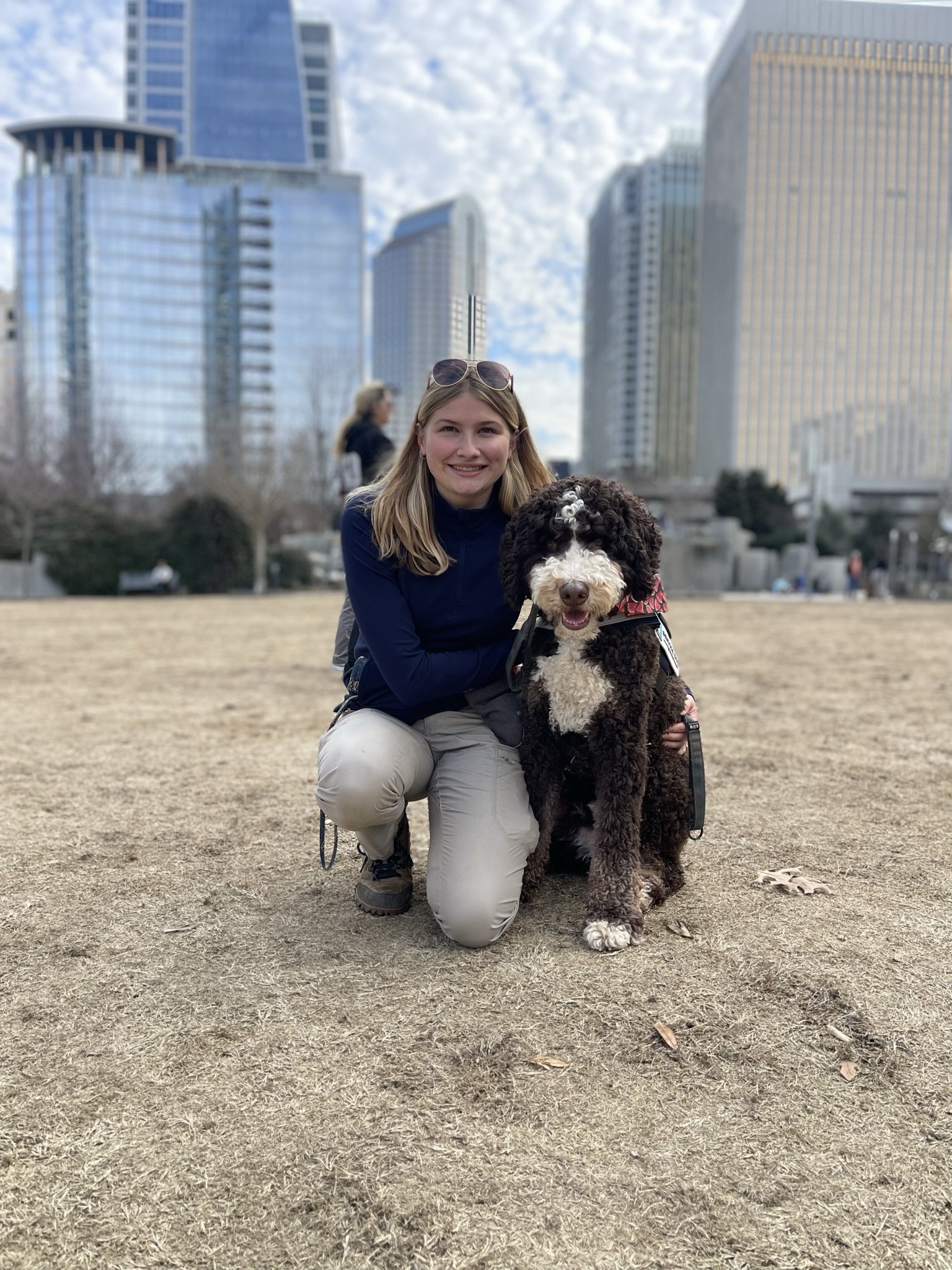 A woman crouching down with a black and white curly-haired dog in an open outdoor area with tall buildings and a partly cloudy sky in the background.