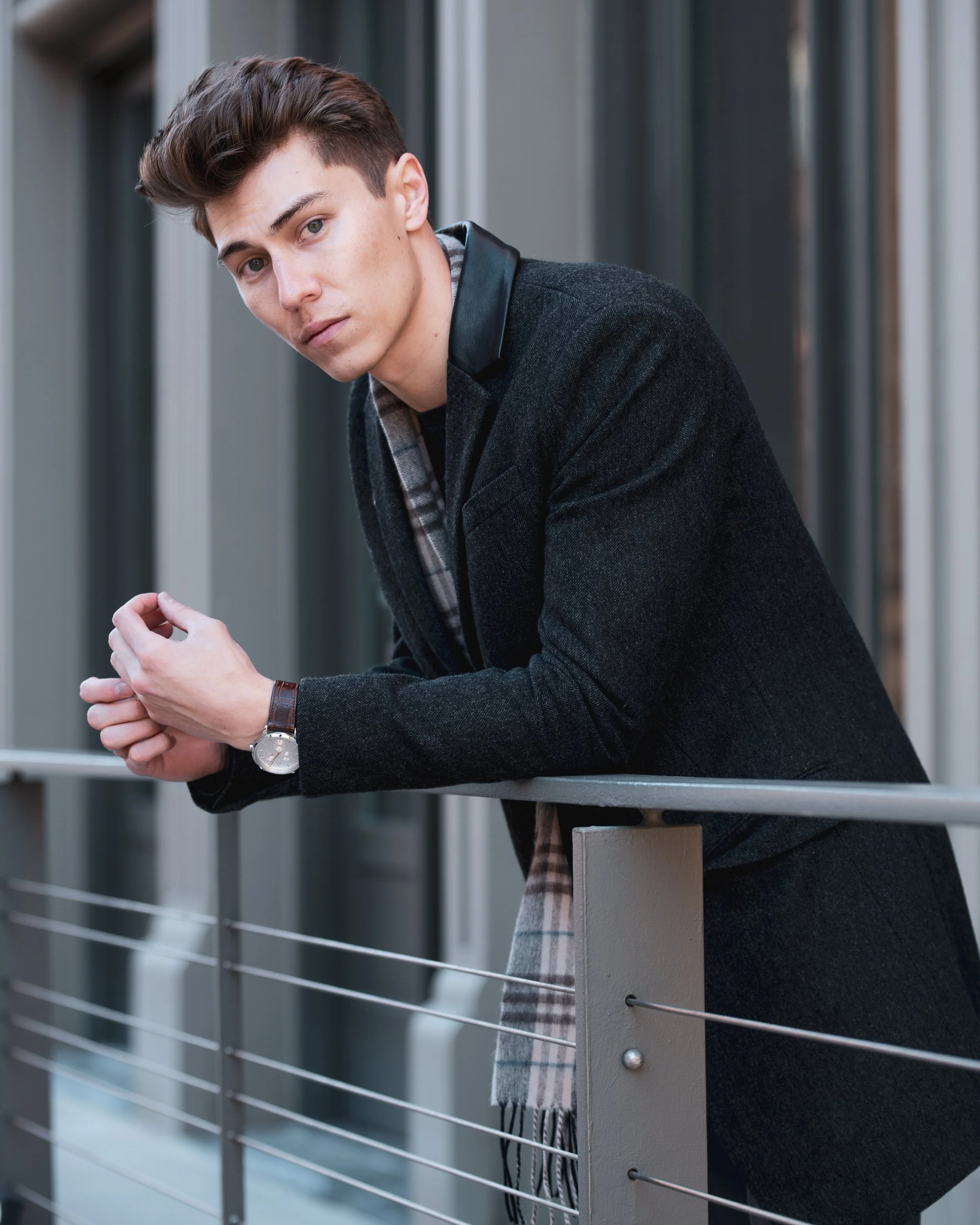A young man with styled brown hair leaning on a metal railing outdoors, wearing a dark coat, a watch, and a plaid scarf, looking at the camera.