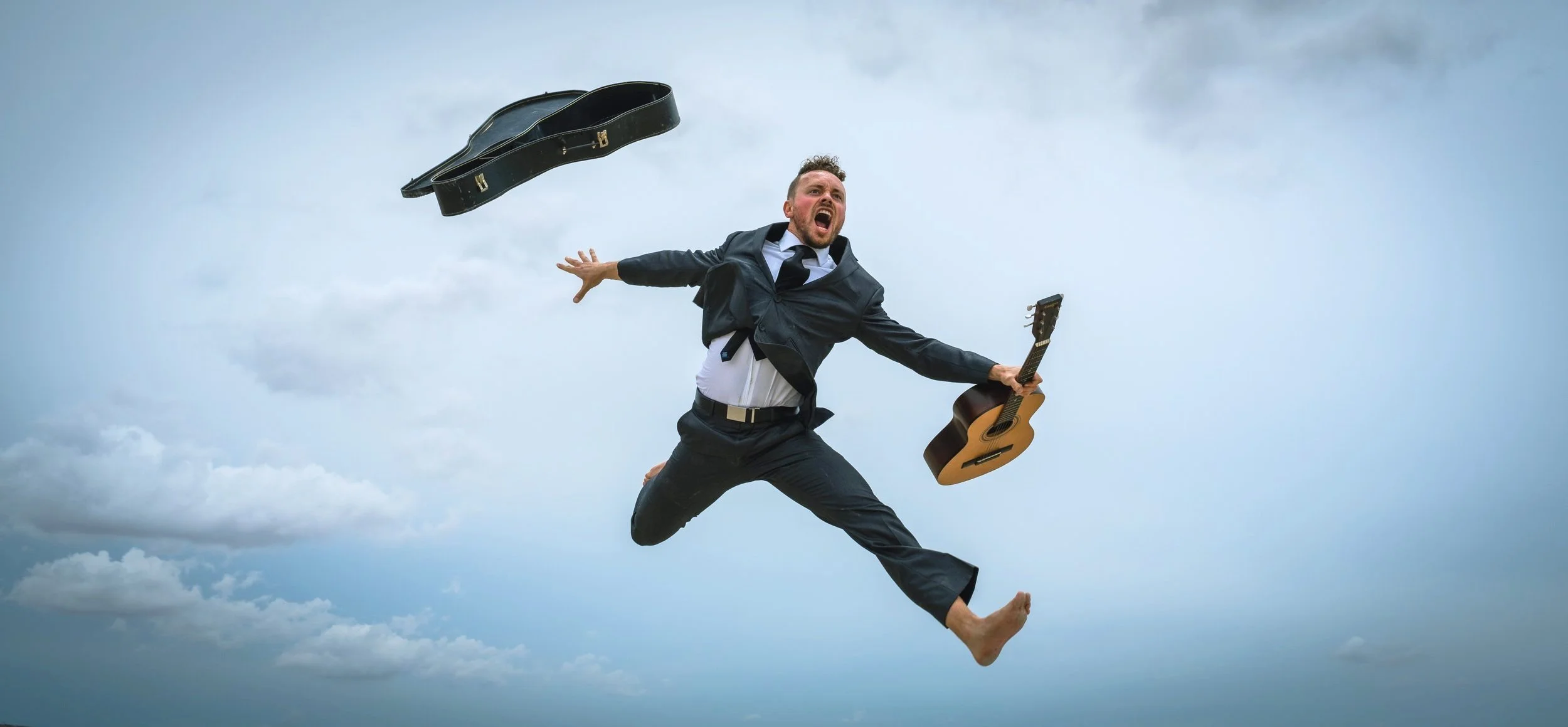 A man in a suit jumping in the air with a guitar in one hand and a guitar case flying nearby against a cloudy sky.