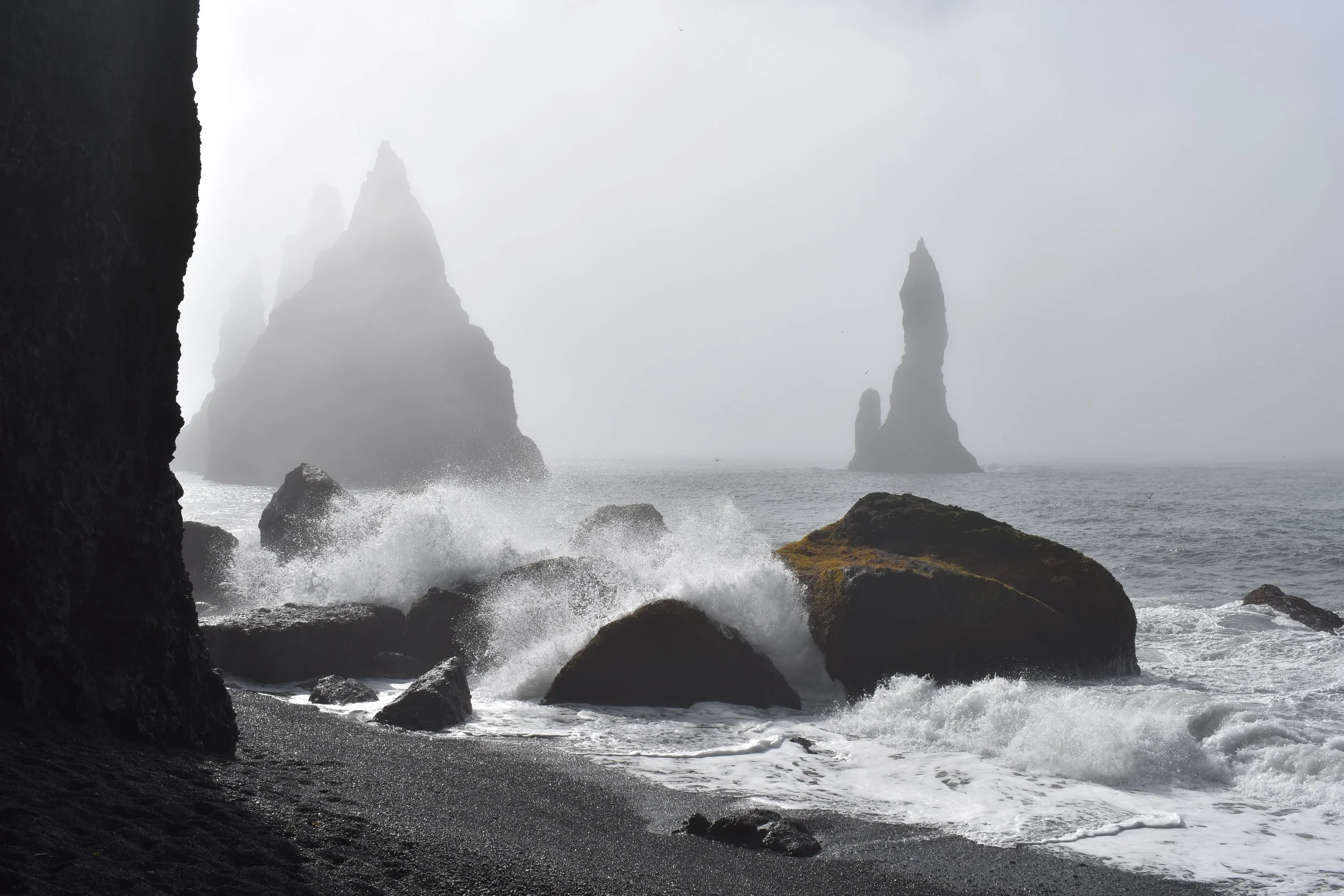 Rocky coast with large sea stacks in the distance, misty and overcast sky, waves crashing against rocks, dark pebbled beach in foreground.