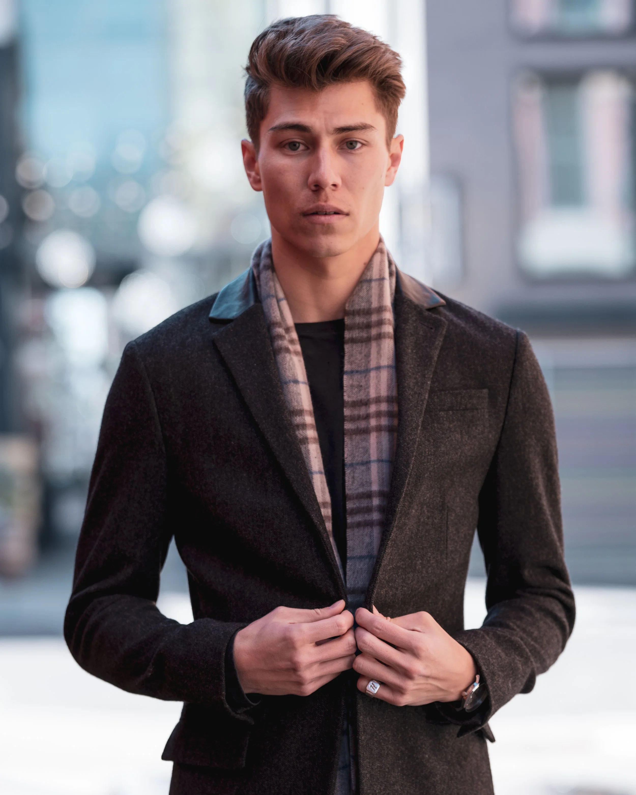 A young man with short brown hair, wearing a dark blazer and a plaid shirt underneath, standing outdoors in front of a building with blurred windows and cityscape in the background.