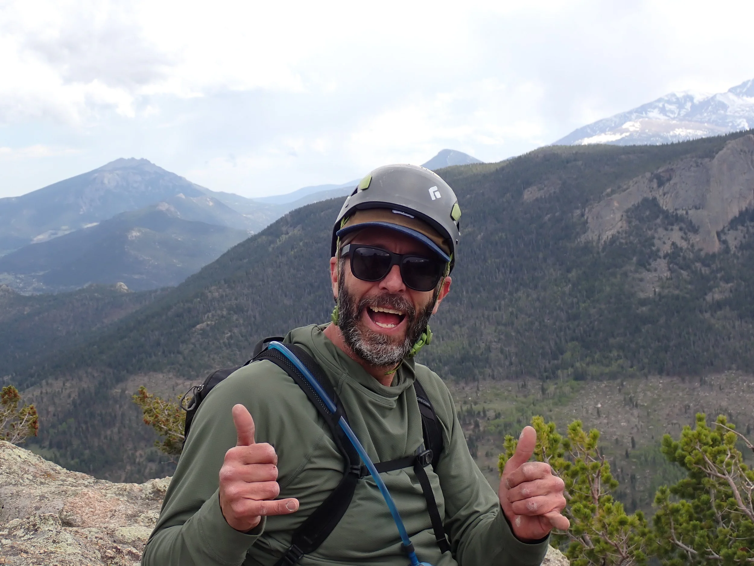A man wearing sunglasses and a helmet, giving two thumbs up on a mountain trail with forested mountains and snow-capped peaks in the background.