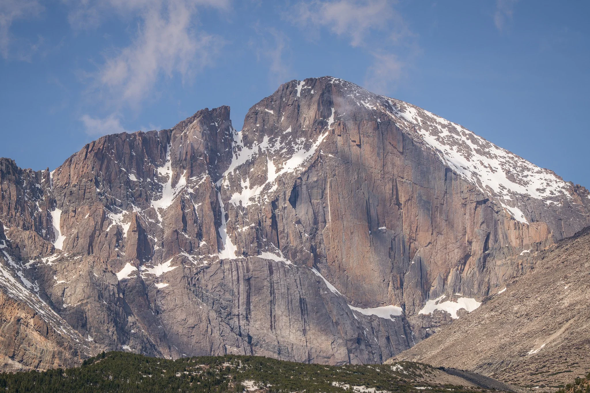 Tall mountain with snow patches and rocky cliffs, green trees at the base, under a blue sky with wispy clouds.