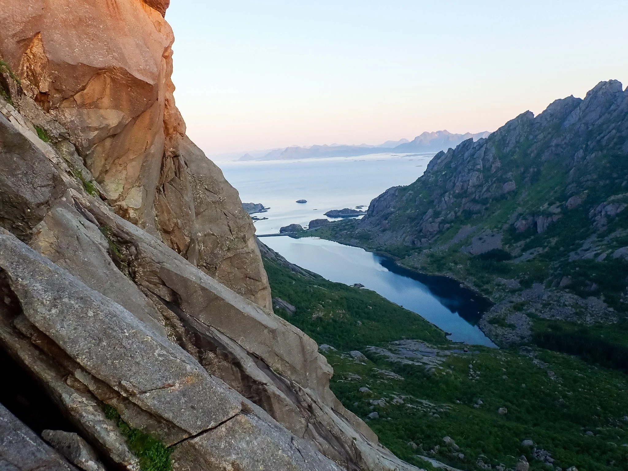 A mountain view with rocky cliffs, a water body winding through green terrain, and distant mountains under a clear sky.