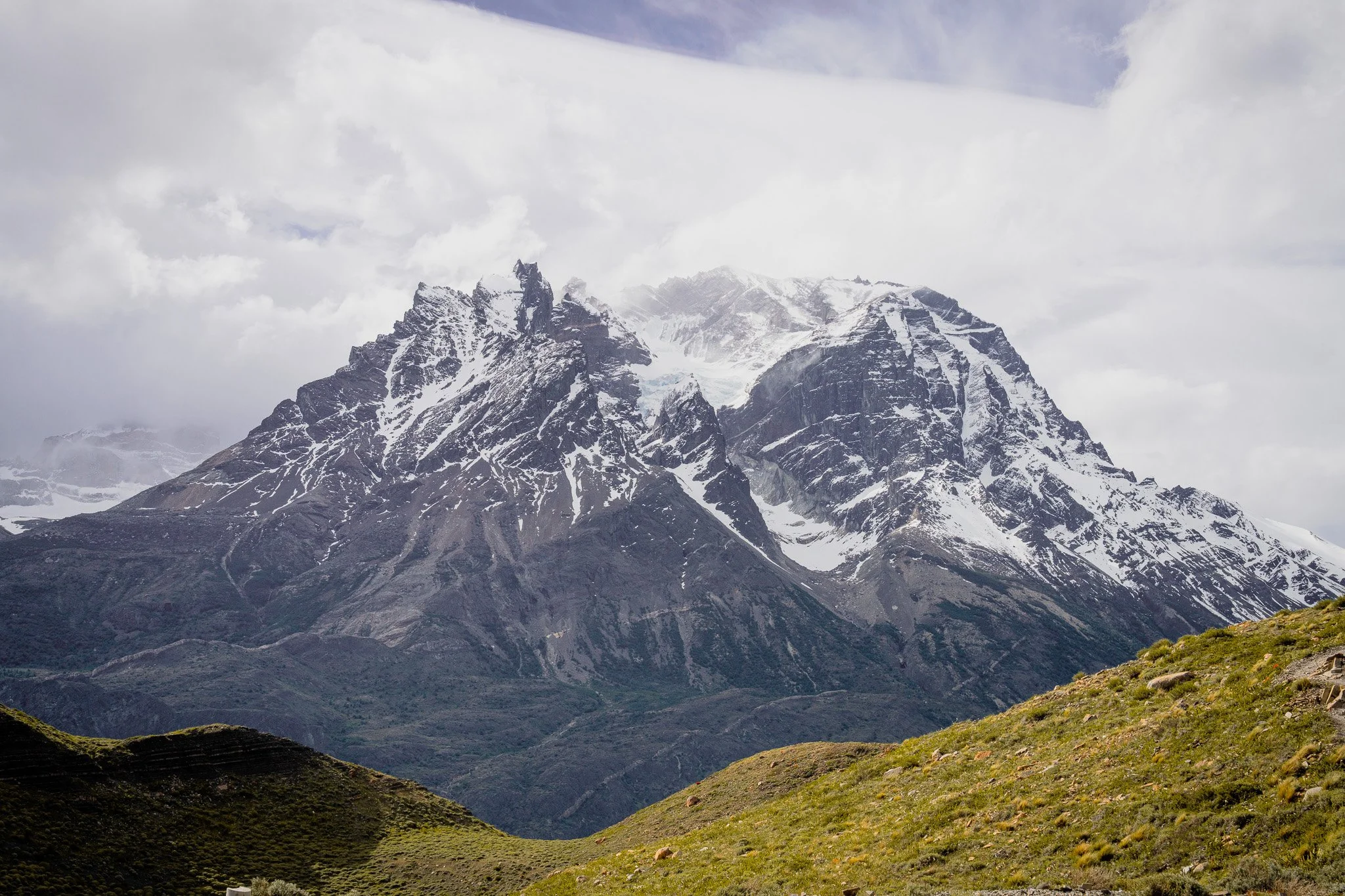 Snow-capped mountains in the background with green grassy hills in the foreground under a cloudy sky.