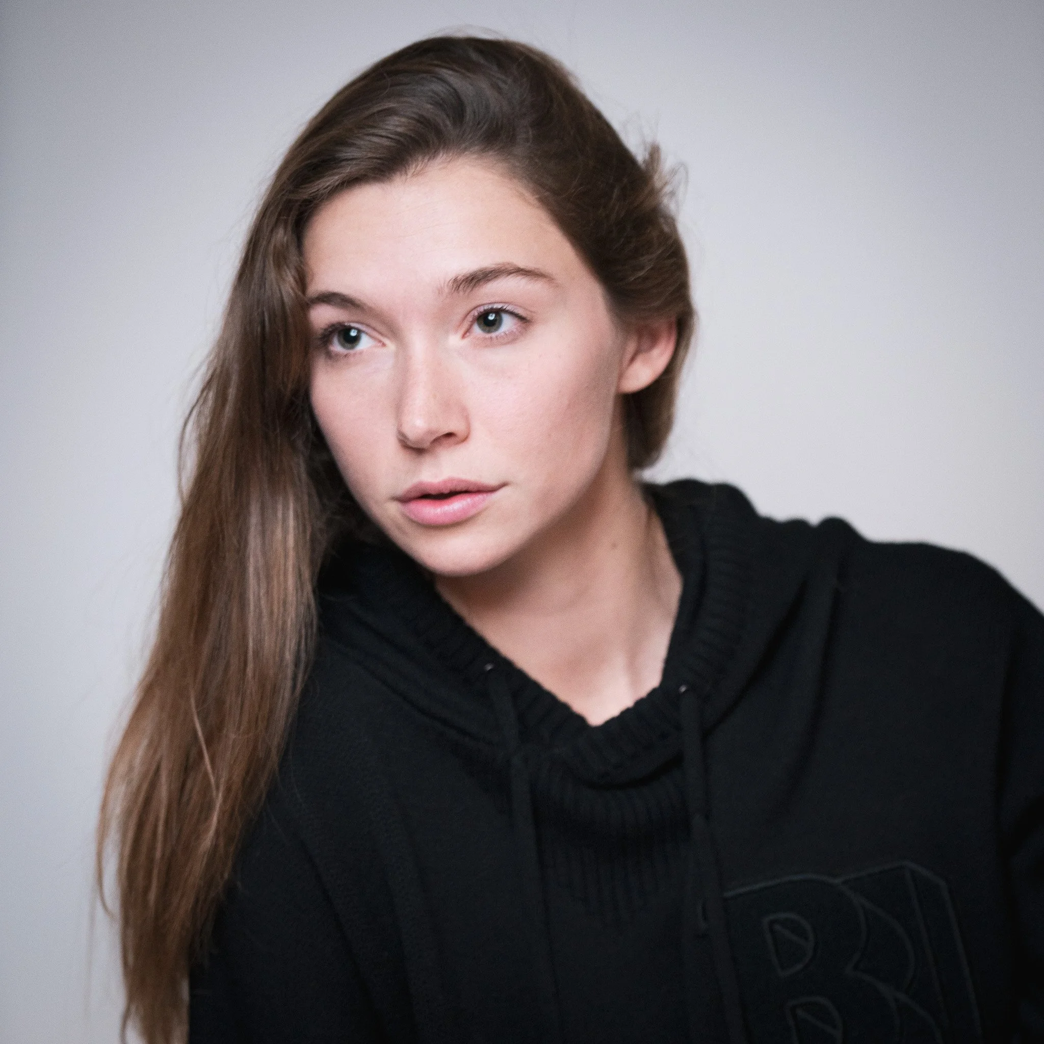 A young woman with long brown hair and fair skin wearing a black hoodie, looking slightly to her left with a neutral expression.