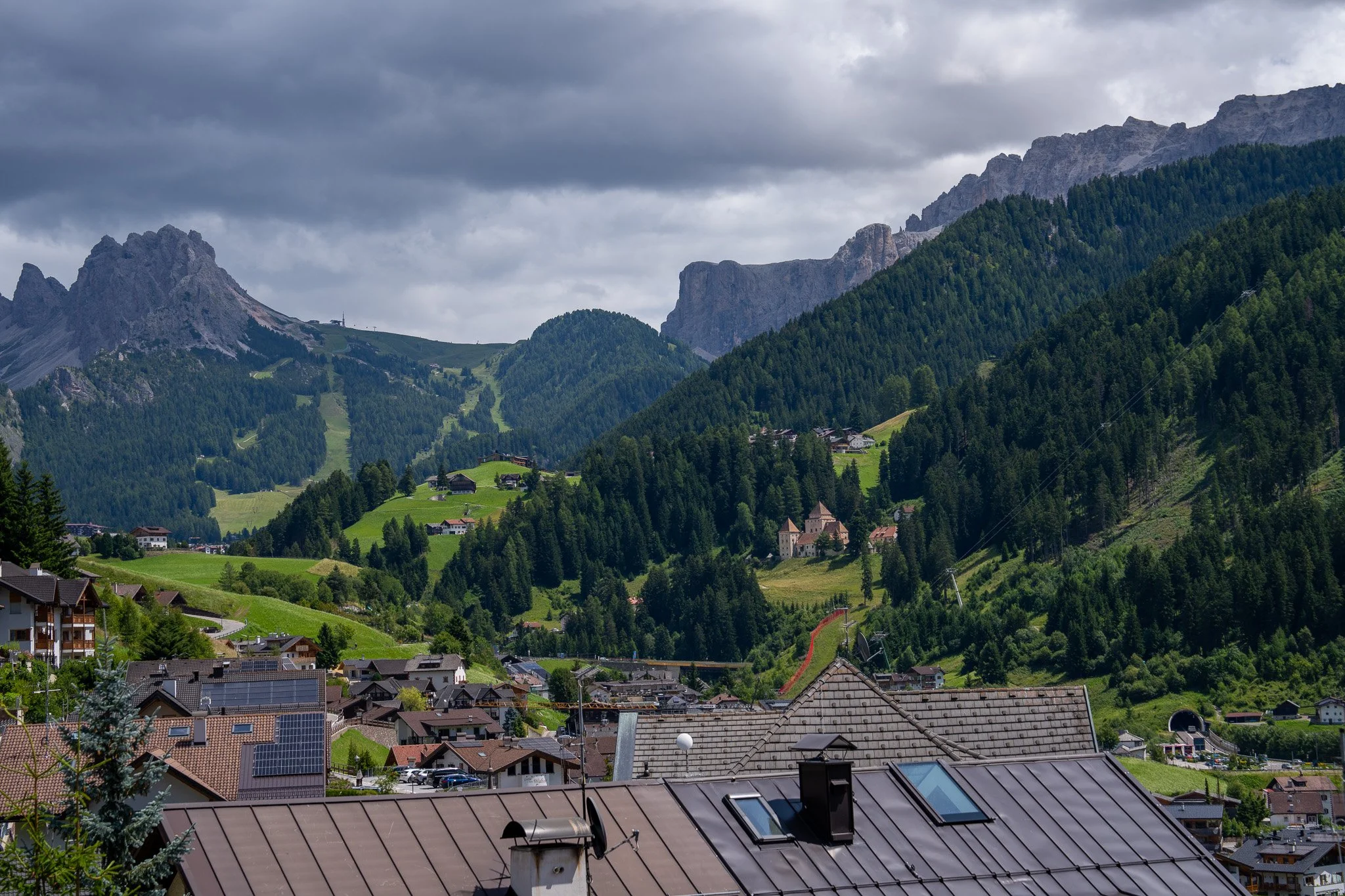 Mountain village with rooftops and solar panels, surrounded by lush green hills and dense forest, with tall mountains in the background under a cloudy sky.