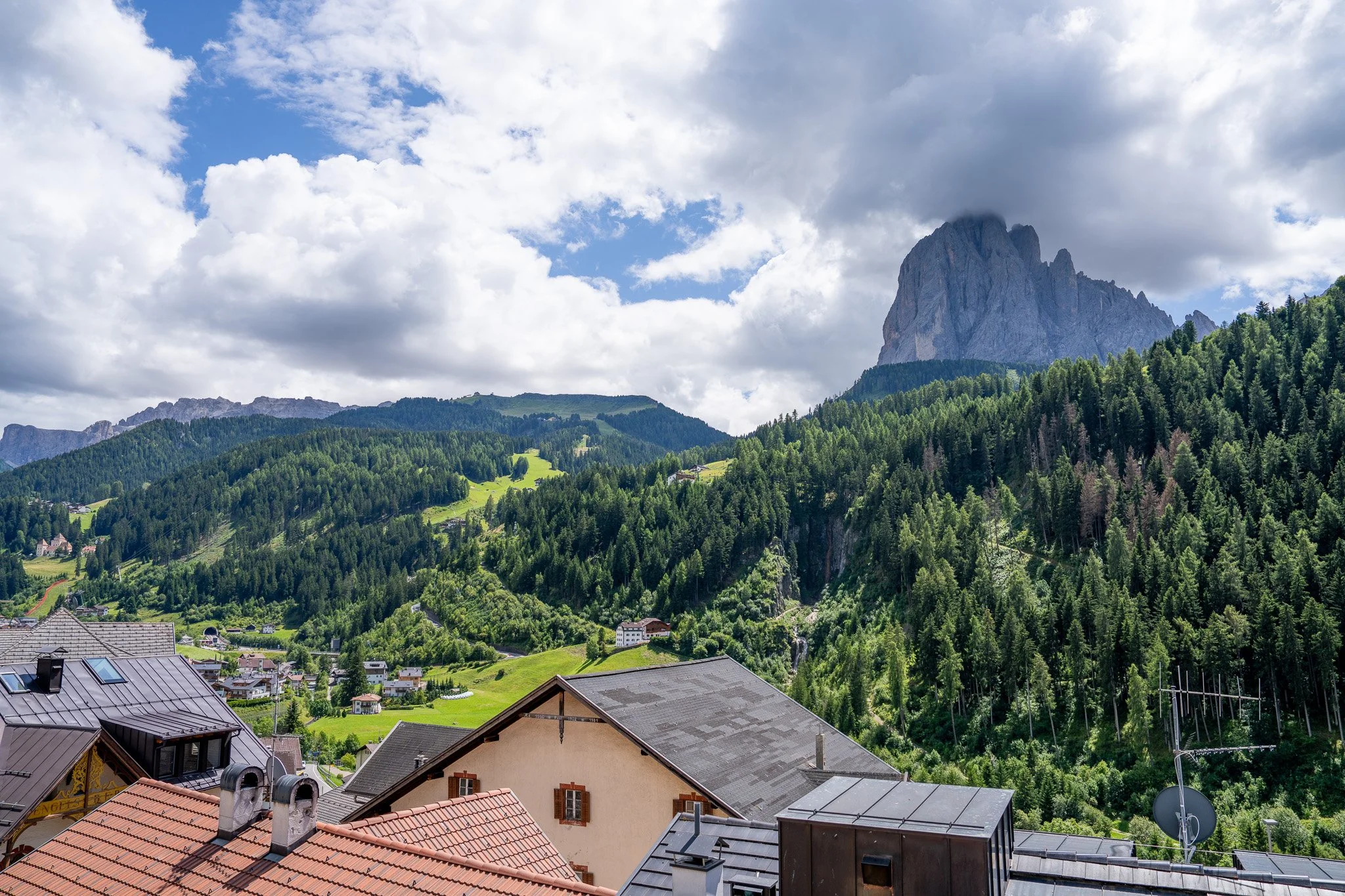 View of a mountain village with rooftops in the foreground, surrounded by green forests and a large mountain with a cloud-covered peak in the background.