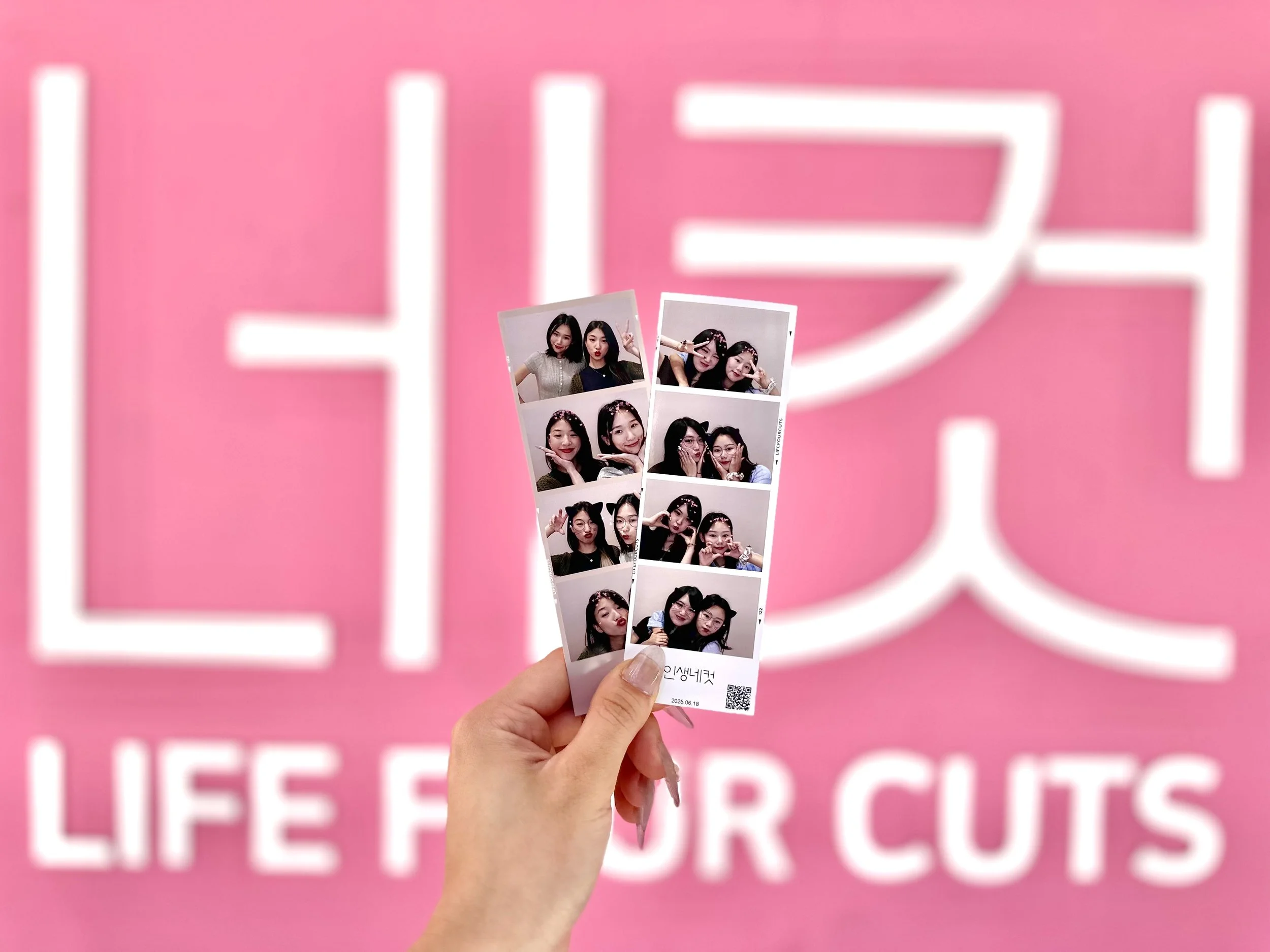 A hand holding printed Life4Cuts photo strips in front of a pink Life4Cuts wall.