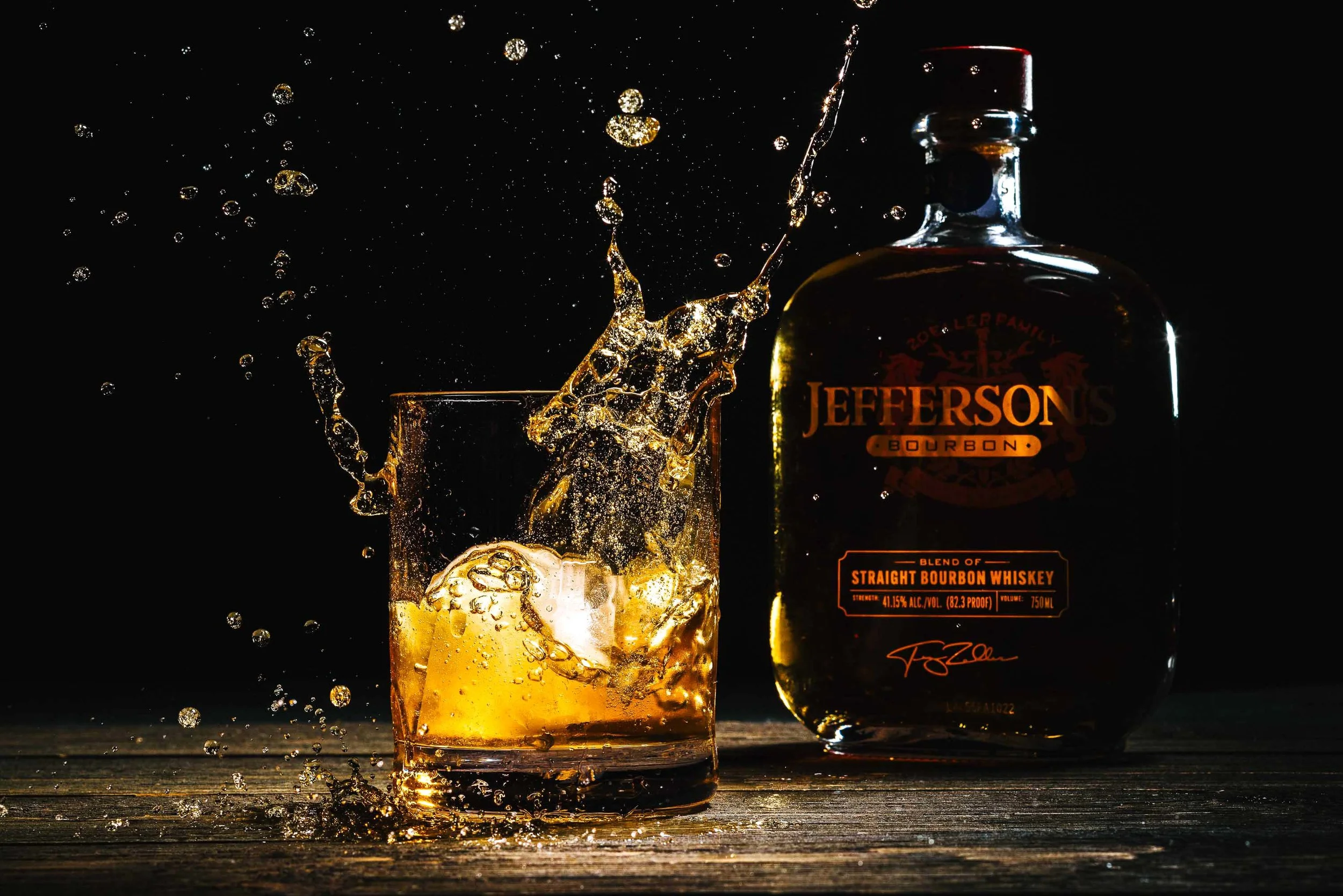 Product photo of an ice cube landing in a glass of whiskey with a bottle of whiskey next to the glass
