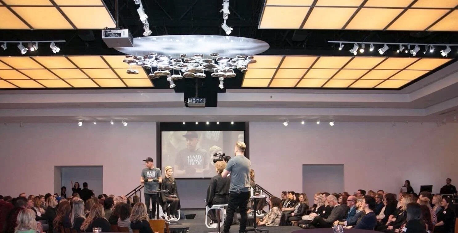 Hairdressing workshop with audience watching stylists on stage under modern ceiling lights.