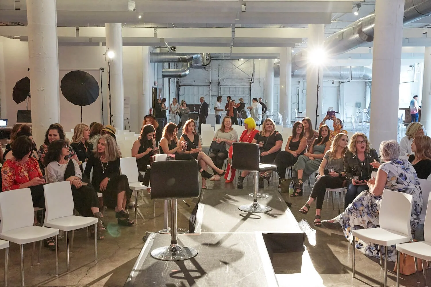 Group of people seated at an indoor event with chairs and a stage in a modern, industrial-style venue.