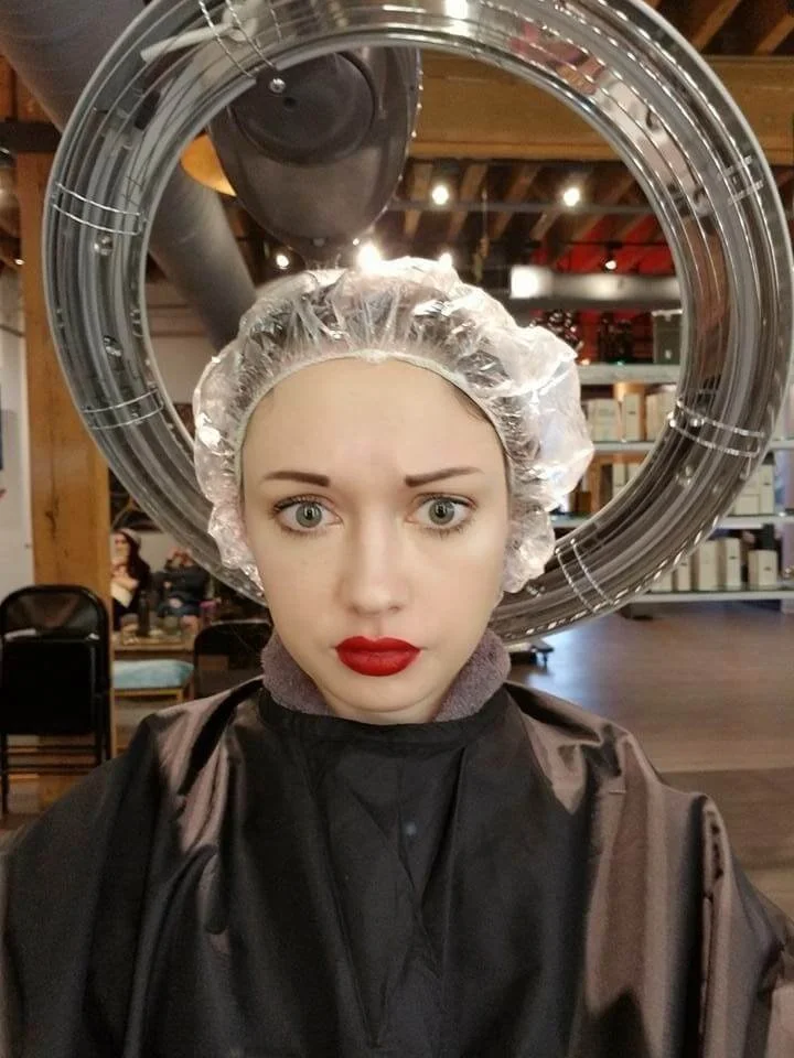 Person wearing a cape and a plastic cap under a hair dryer in a salon.