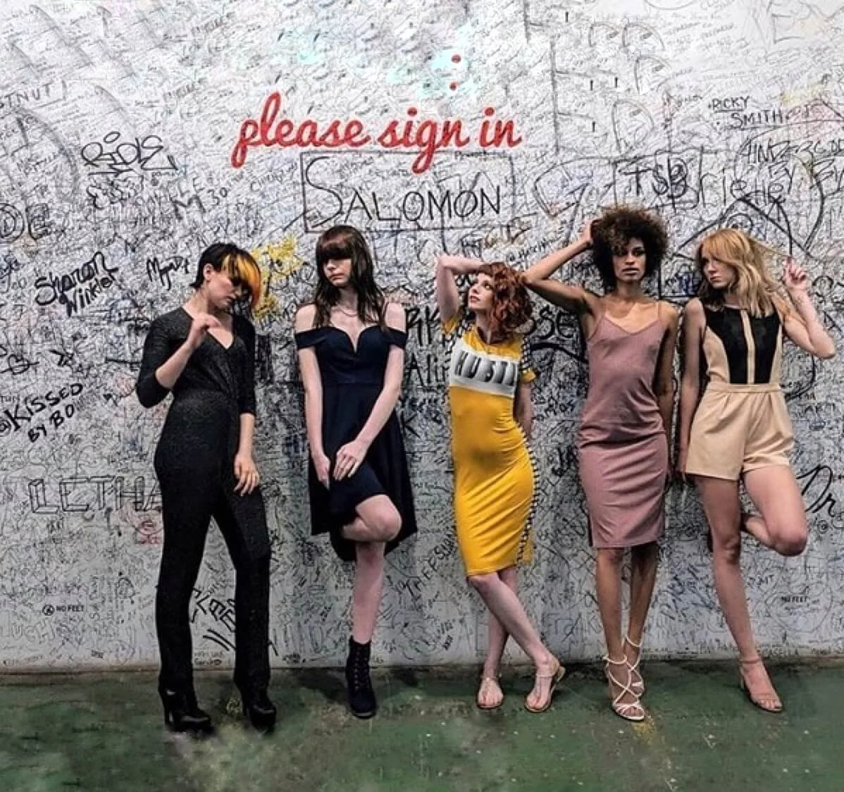 Five women standing against a graffitied wall, each posing in various outfits and styles.