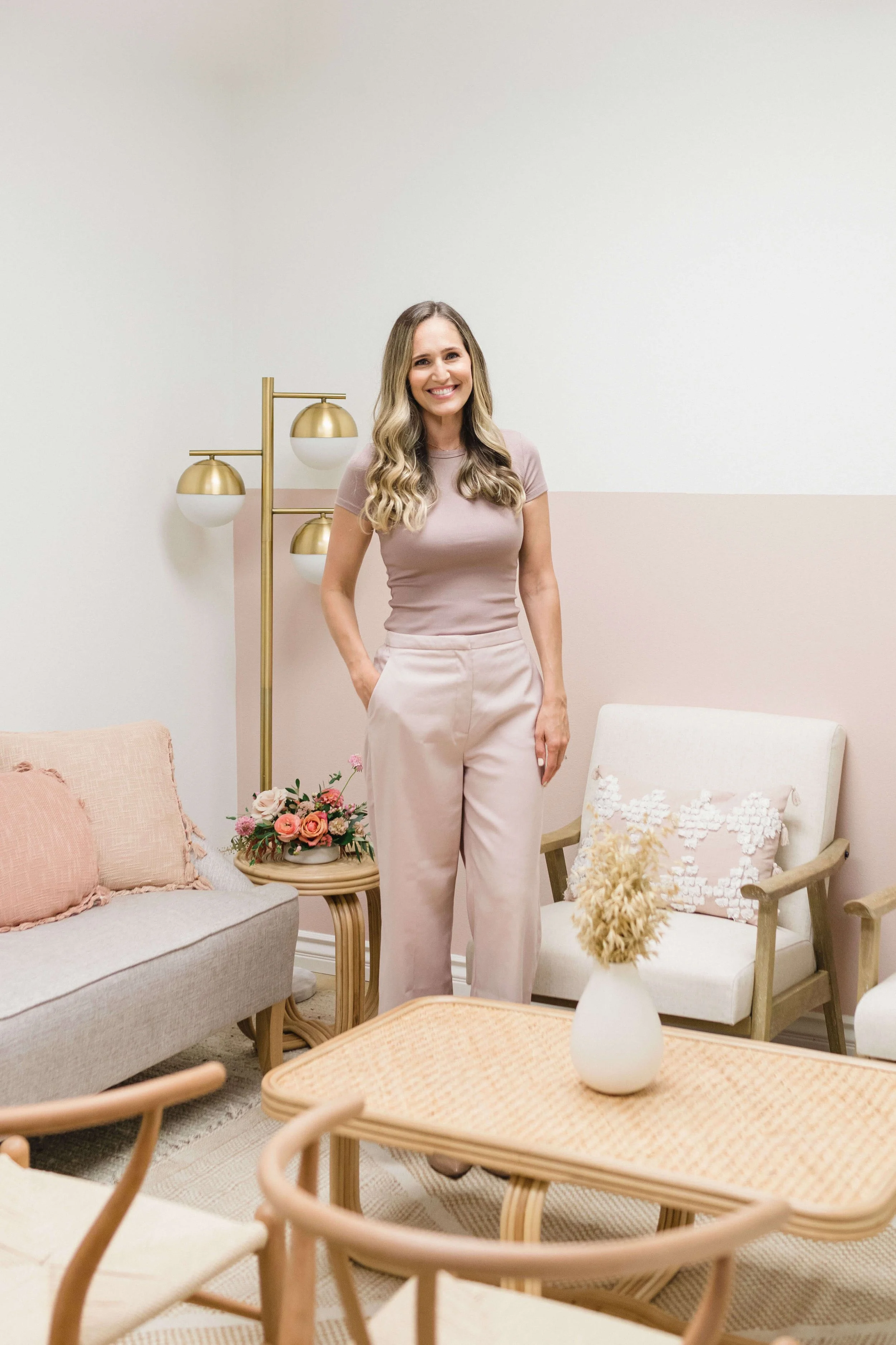 A woman with long blonde hair, wearing a pink t-shirt and light pink pants, standing in a cozy, pastel-decorated therapy for anxiety room with floral arrangements and modern furniture.