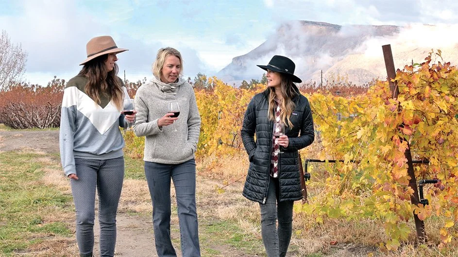 3 women in warm clothing walking through a fall colored vineyard with glasses of wine in hand.