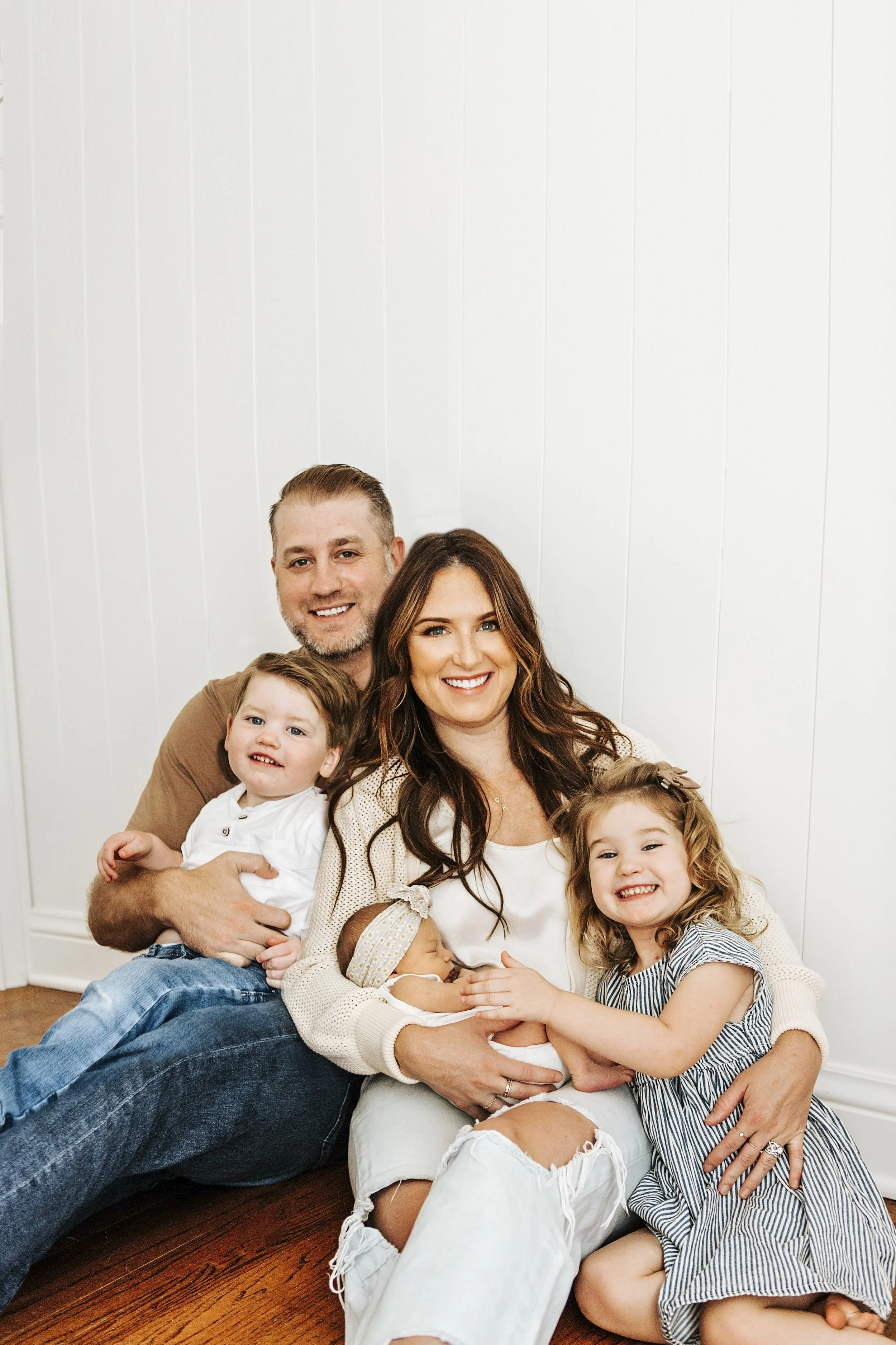 A happy family of five sitting on the wooden floor in front of a white wall, including a father, mother, two young children, and a newborn baby.