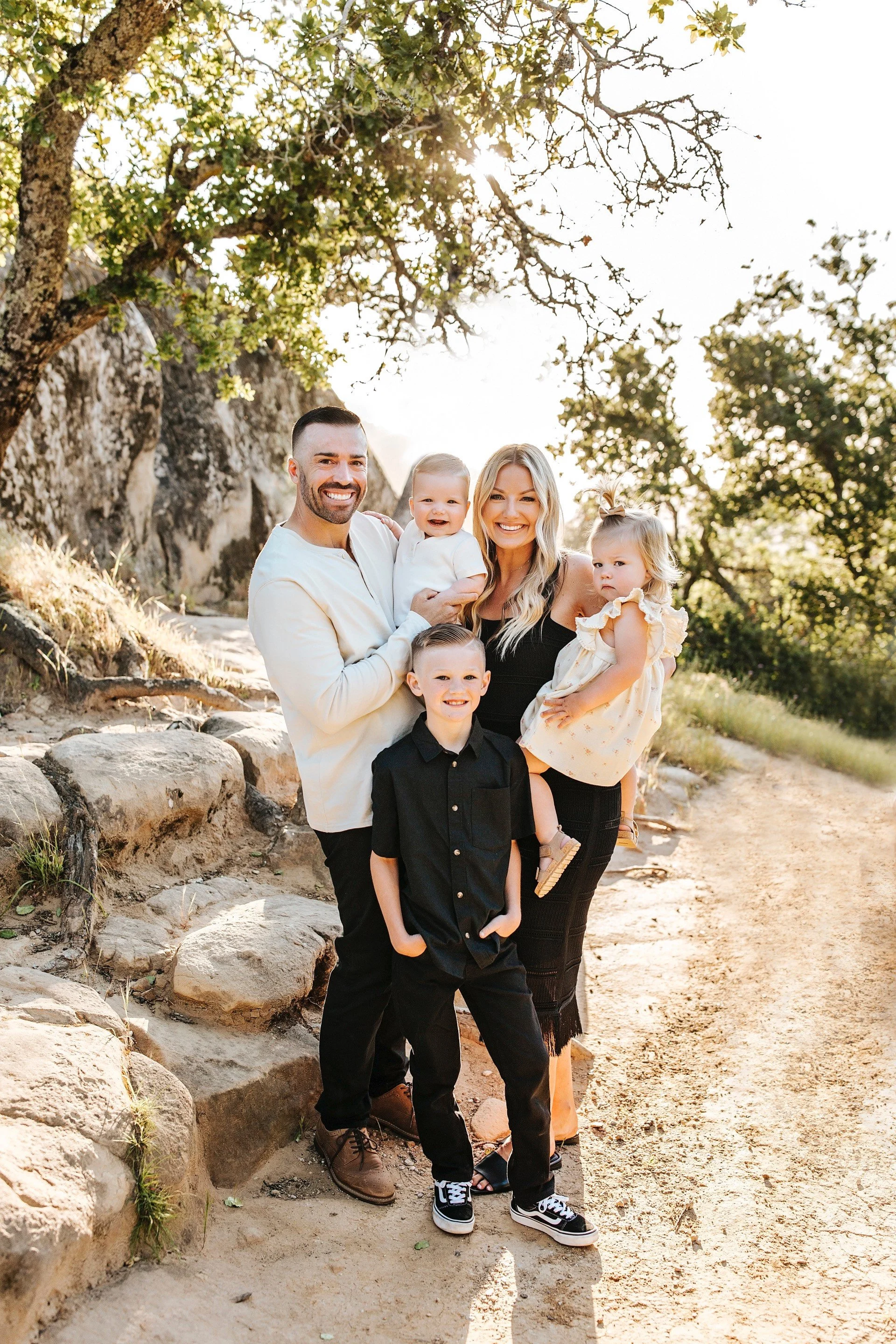 A family of five outdoors, standing on a dirt path near rocks and trees, smiling and posing for a photo in warm sunlight.