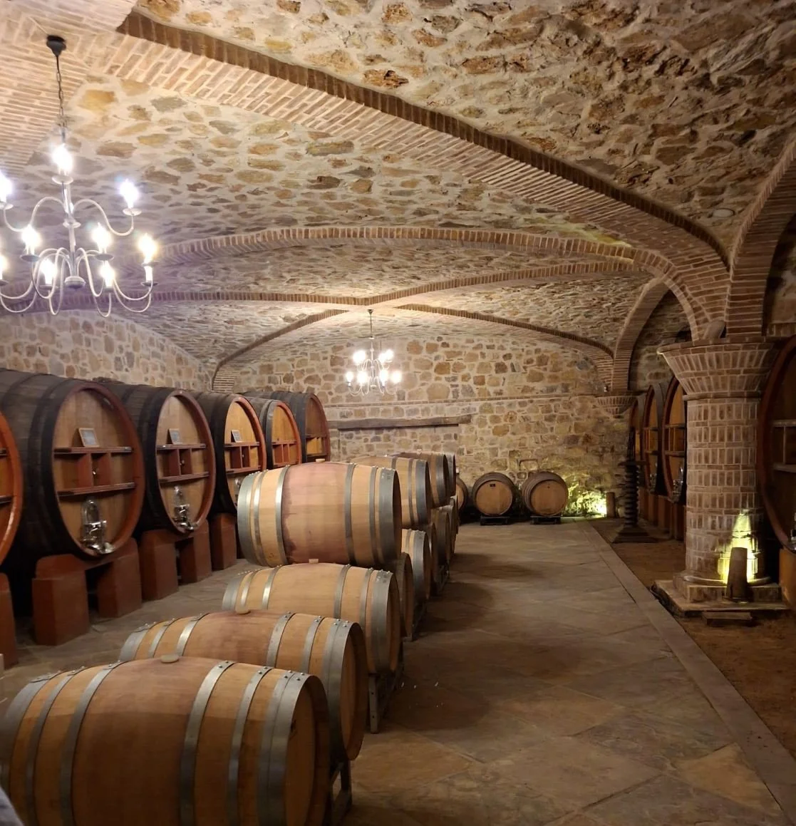 A majestic brick and stone vaulted wine cellar at Chateau Rasque in Provence, featuring rows of large oak tuns and aging barrels under lit chandeliers.