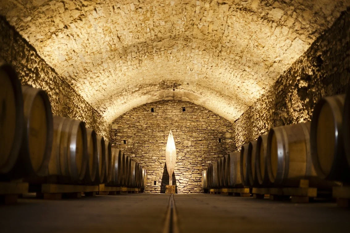 A symmetrically lit historic stone barrel cellar featuring rows of large oak aging casks and a central stone sculpture.