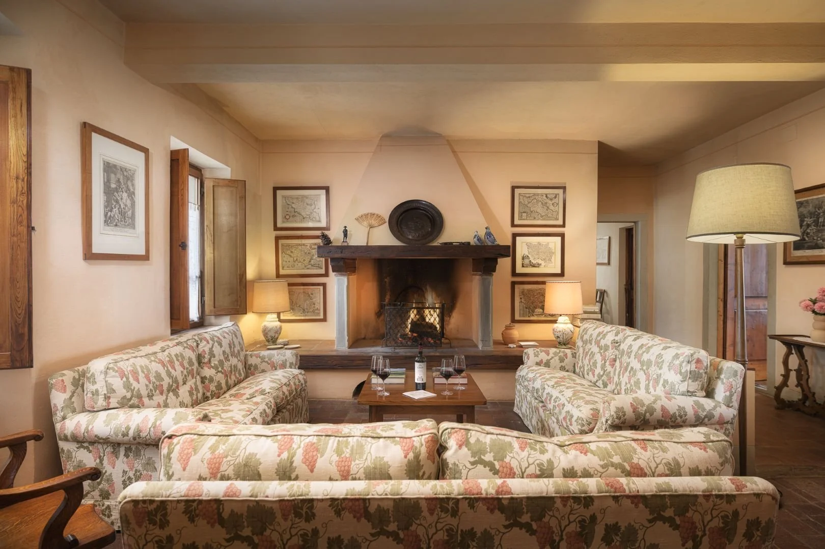 A charming, rustic living room inside a historic vineyard villa featuring a roaring fireplace, comfortable floral sofas, and wine glasses on a coffee table.