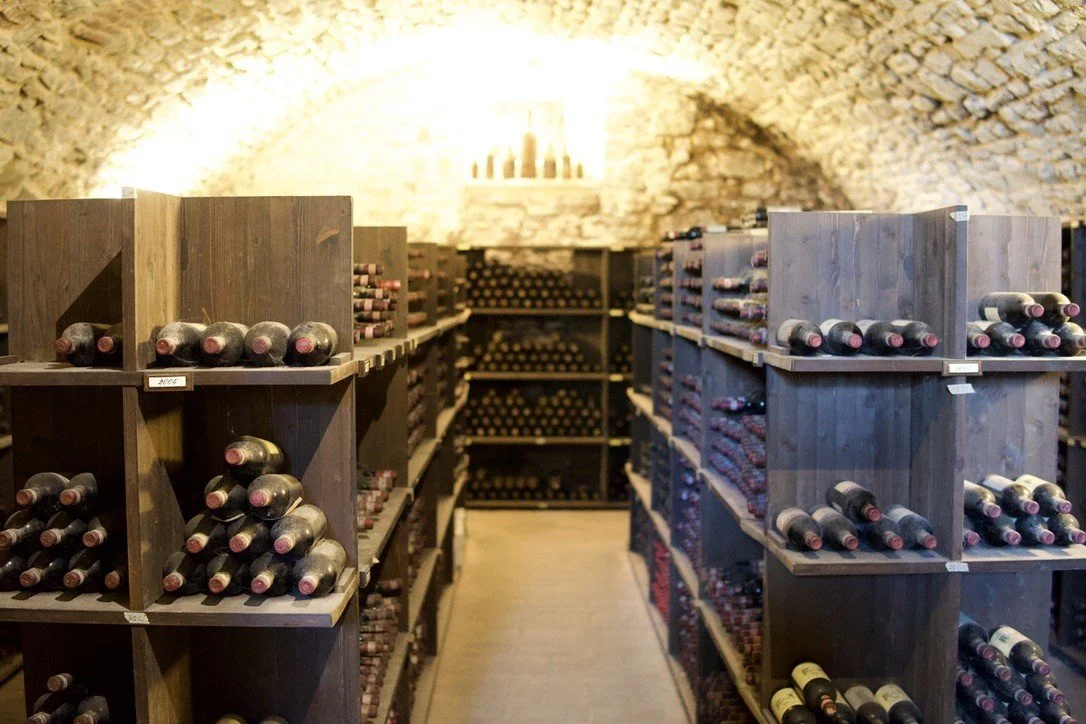 An atmospheric, historic stone wine cellar with rustic wooden shelves stacked with aging vintage wine bottles in Tuscany.