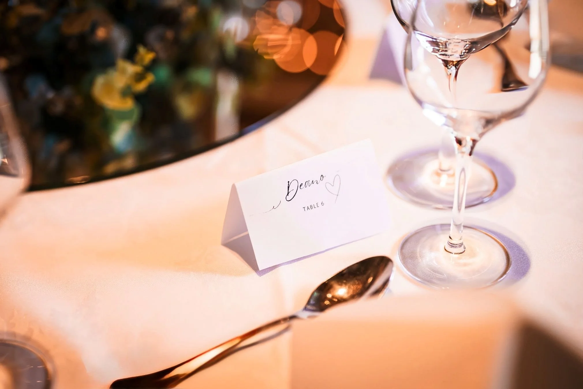 A close-up of an elegant wedding table place setting with a custom name card and fine wine glass under warm reception lighting.
