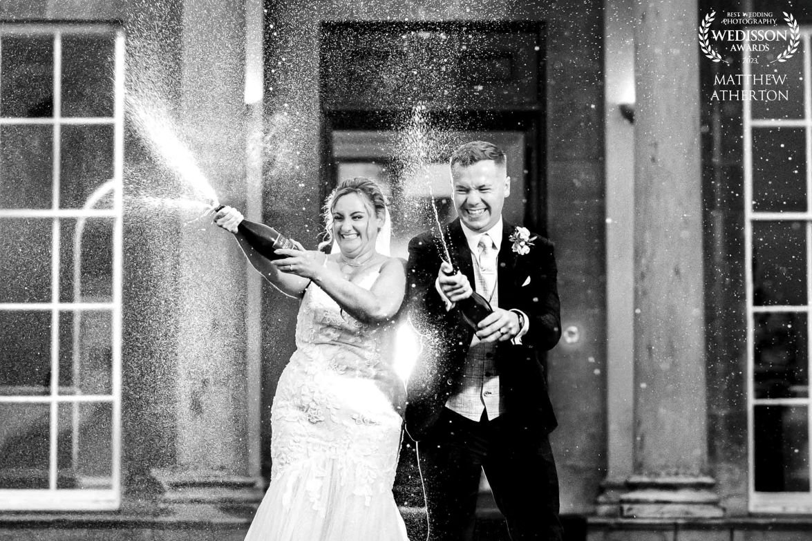 A bride and groom joyfully spraying bottles of champagne outside a grand wedding venue in the North West, captured by Matthew Atherton Photography.
