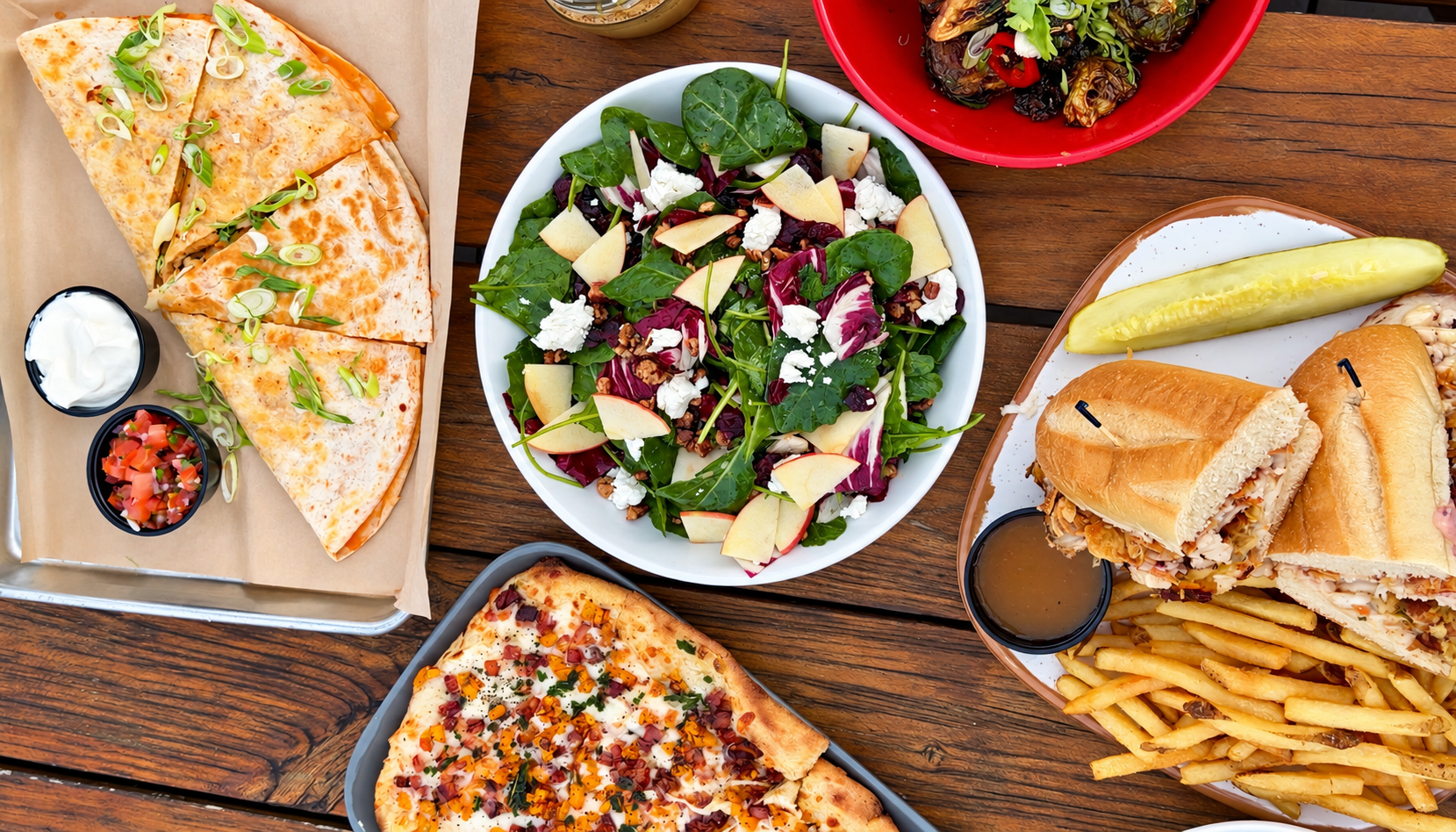 A table with various foods including a veggie salad, a pizza, a sub sandwich with fries and a pickle, a cheese quesadilla, and a plate of roasted vegetables.
