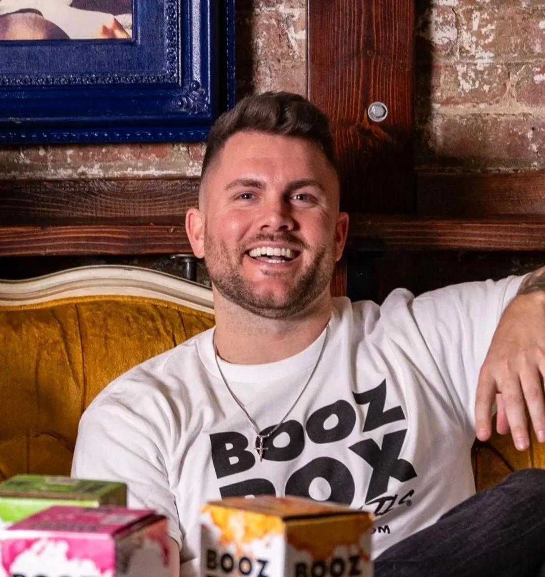 A smiling man with short dark hair and a beard, wearing a white t-shirt with black text, sitting on a yellow upholstered chair in a rustic setting with brick and wood walls, with various colorful boxes of tea or snacks in front of him.