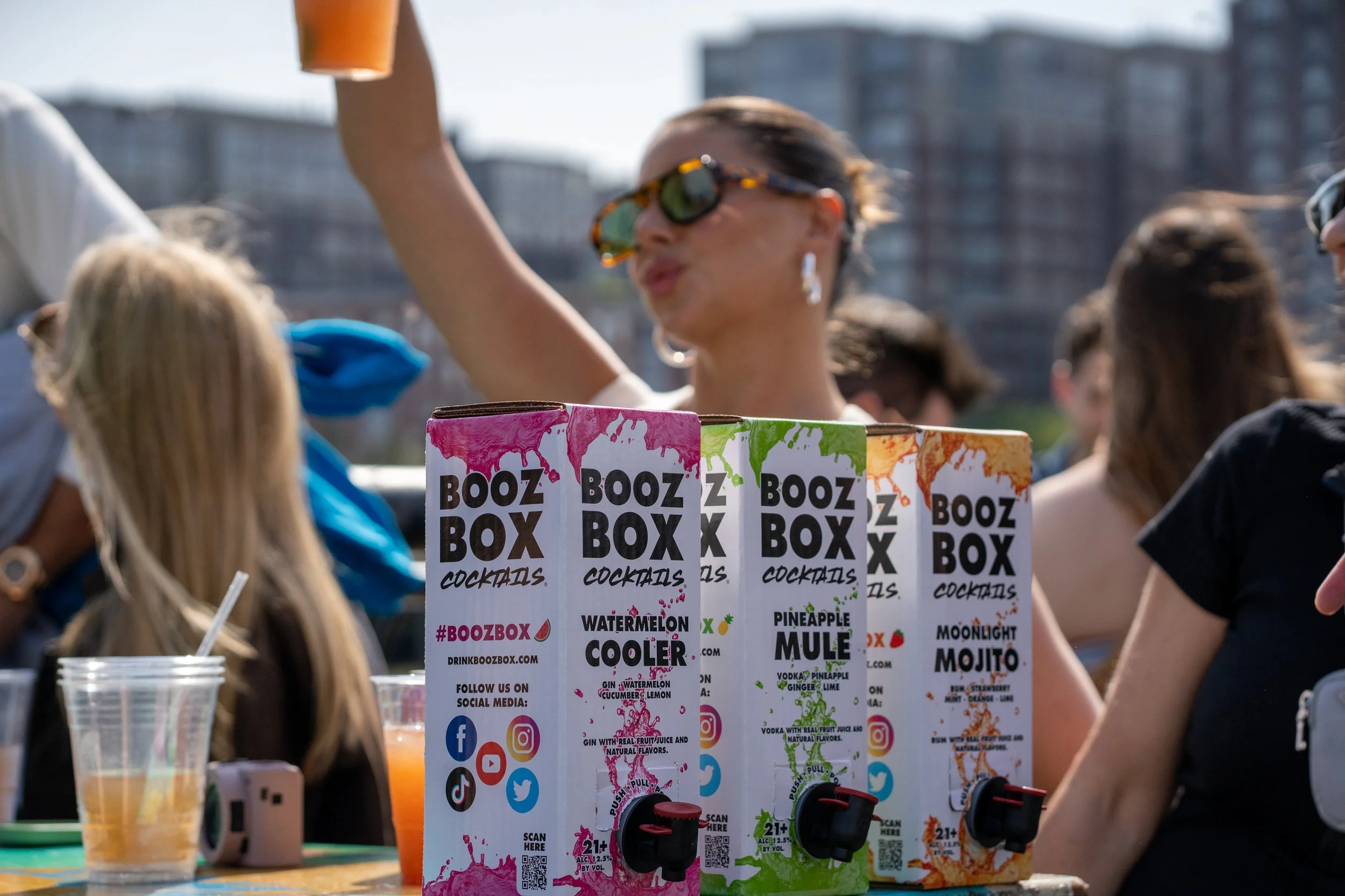 Booz Box cocktails on a table at an outdoor event with people in the background.