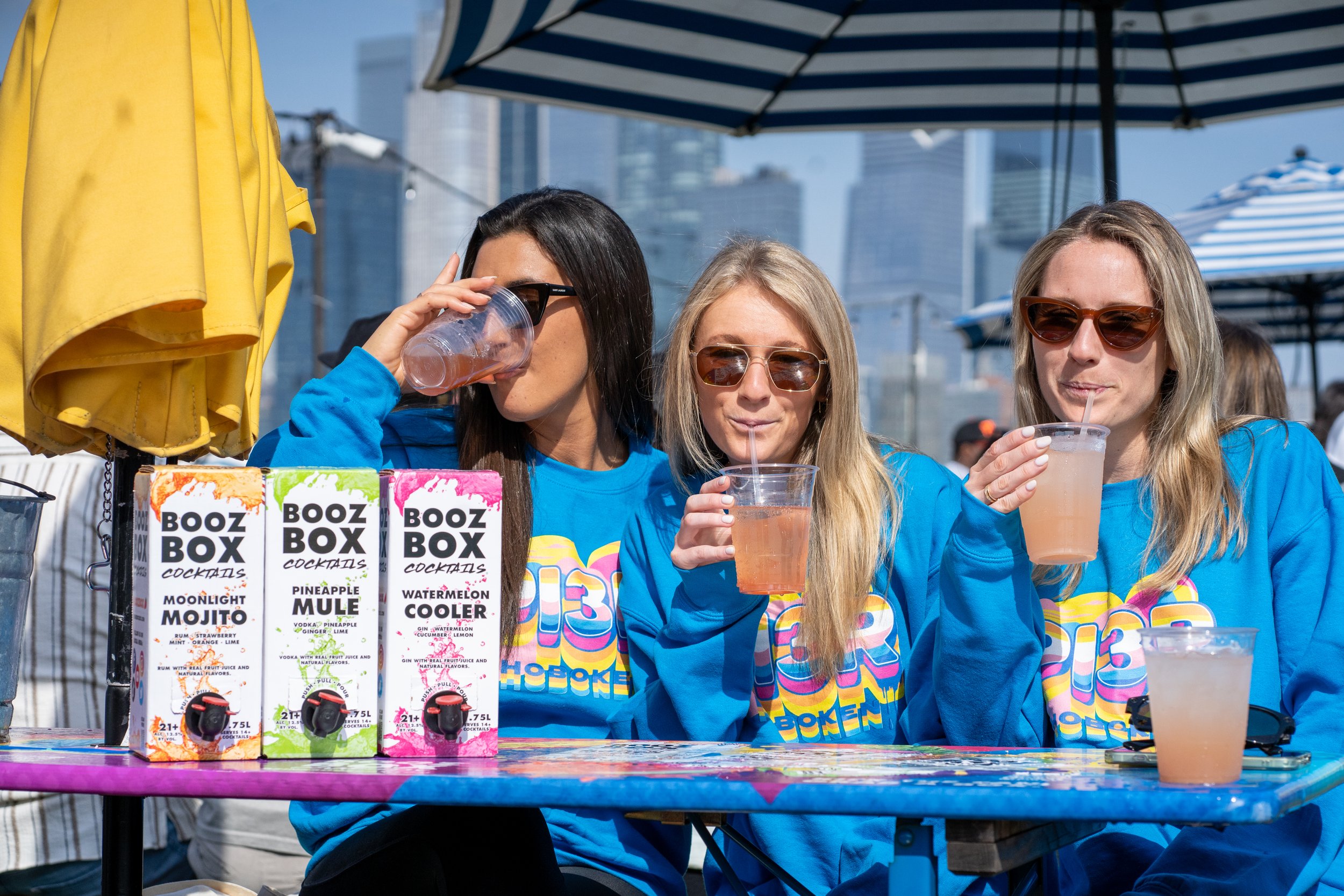 Three women sitting at an outdoor table, drinking pink beverages through straws, with a city skyline and umbrellas in the background, wearing blue shirts and sunglasses.