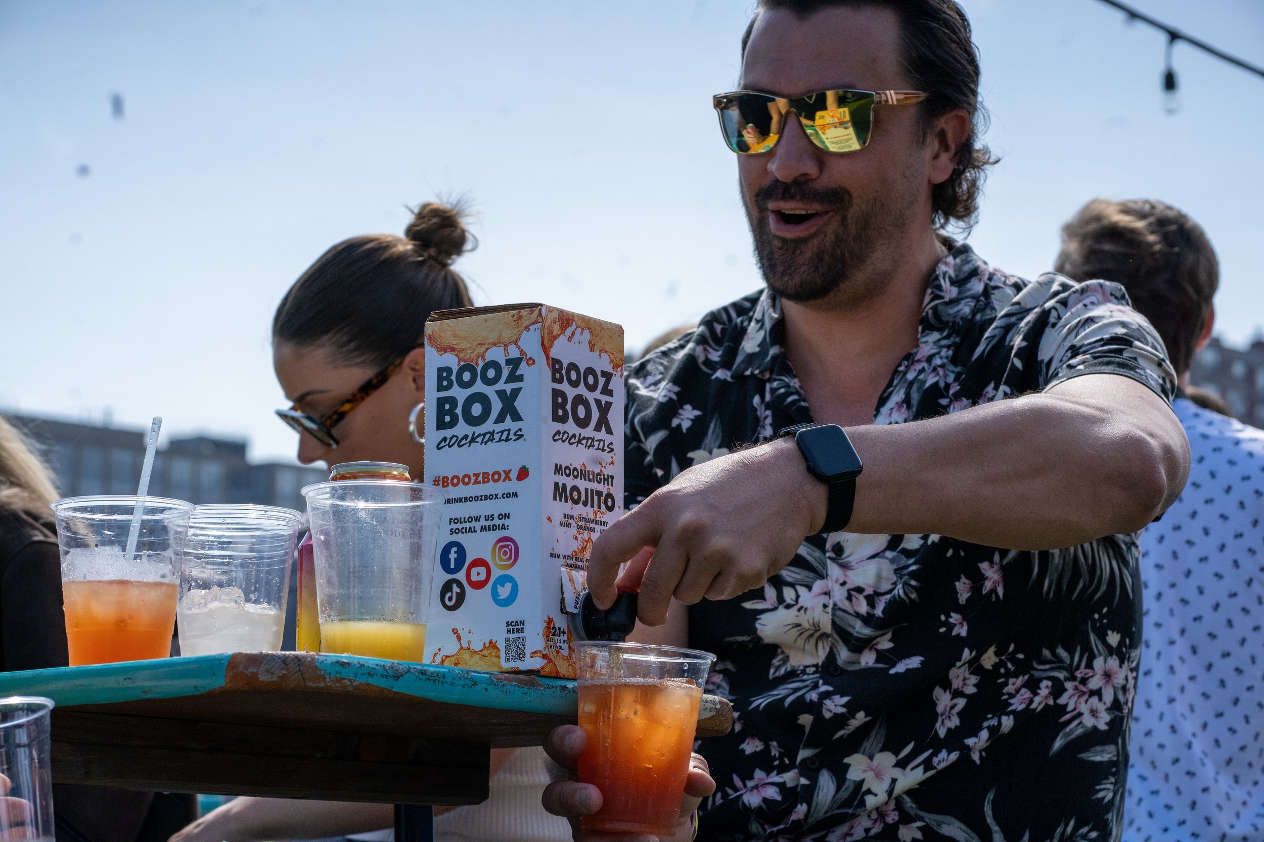 A man wearing sunglasses and a floral shirt is pouring a drink at an outdoor party. There are drinks and a menu on the table.