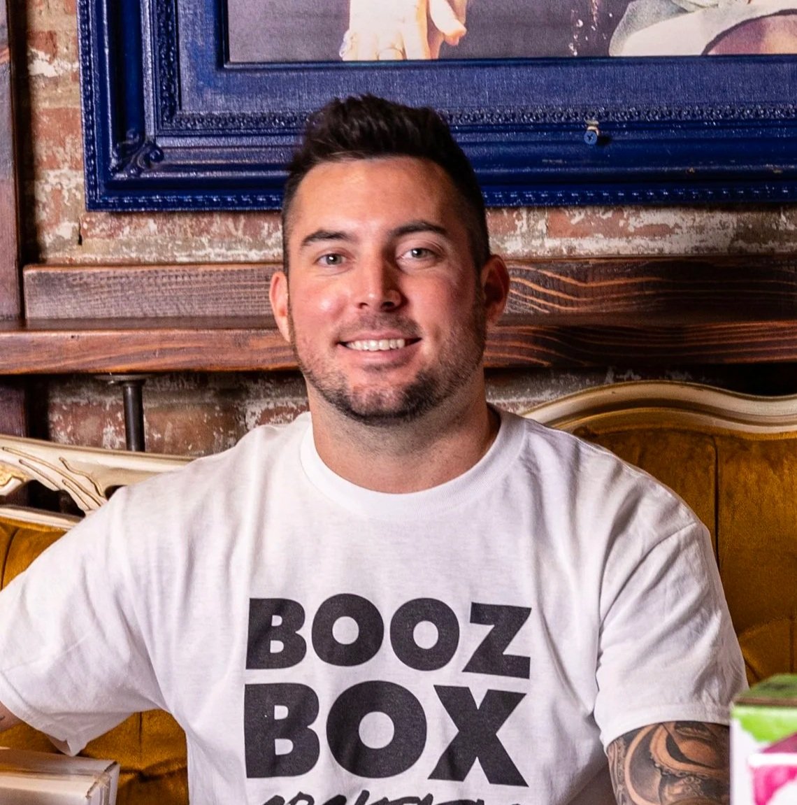 A man with short dark hair, a beard, and a white T-shirt with bold black letters smiling while sitting at a table in a restaurant.